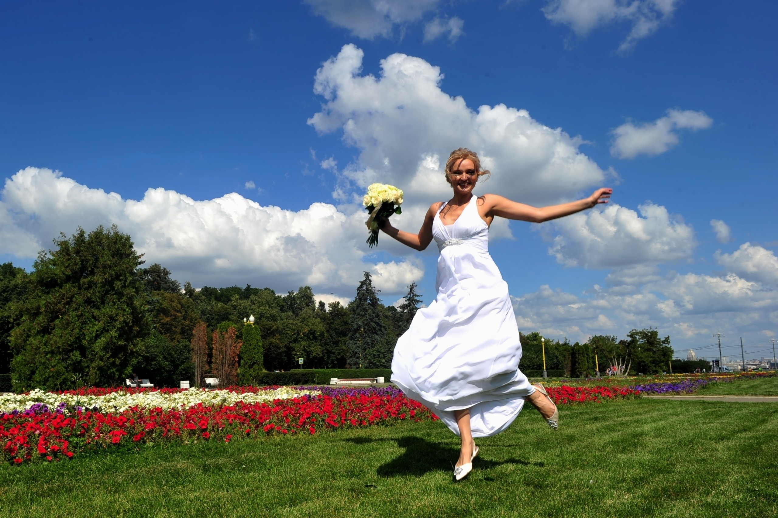 Свадьба на Воробьёвых Горах рядом с МГУ, невеста радостно бежит по зеленой лужайке со свадебным букетом из кремовых роз в руке, на заднем плане ярко-синее небо, красные и белые цветы и изумрудная зелень лужайки