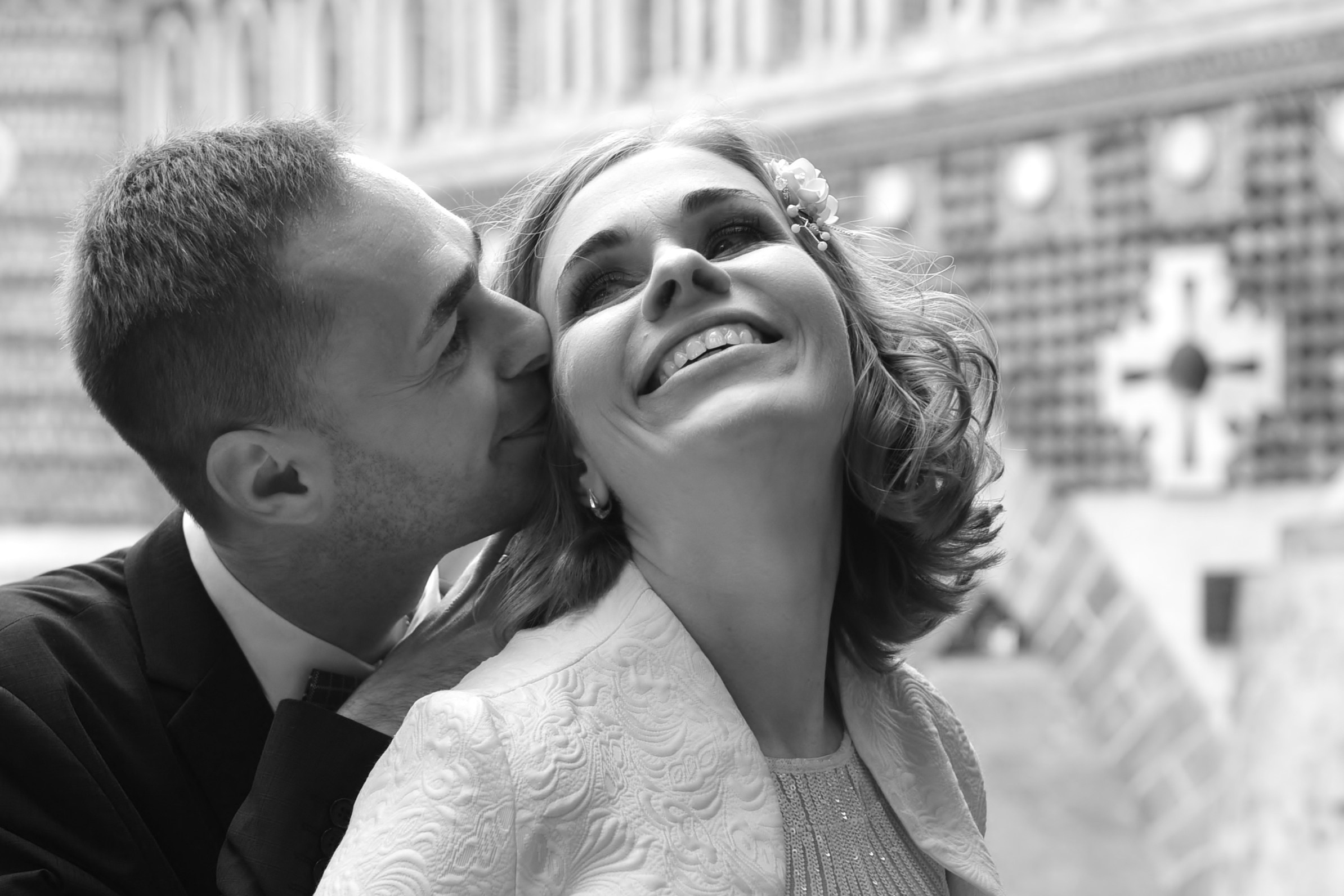 Свадьба в Царицыно хмурым прохладным летним днём, жених целует невесту в щеку на фоне кирпичного моста, чёрно-белая фотография