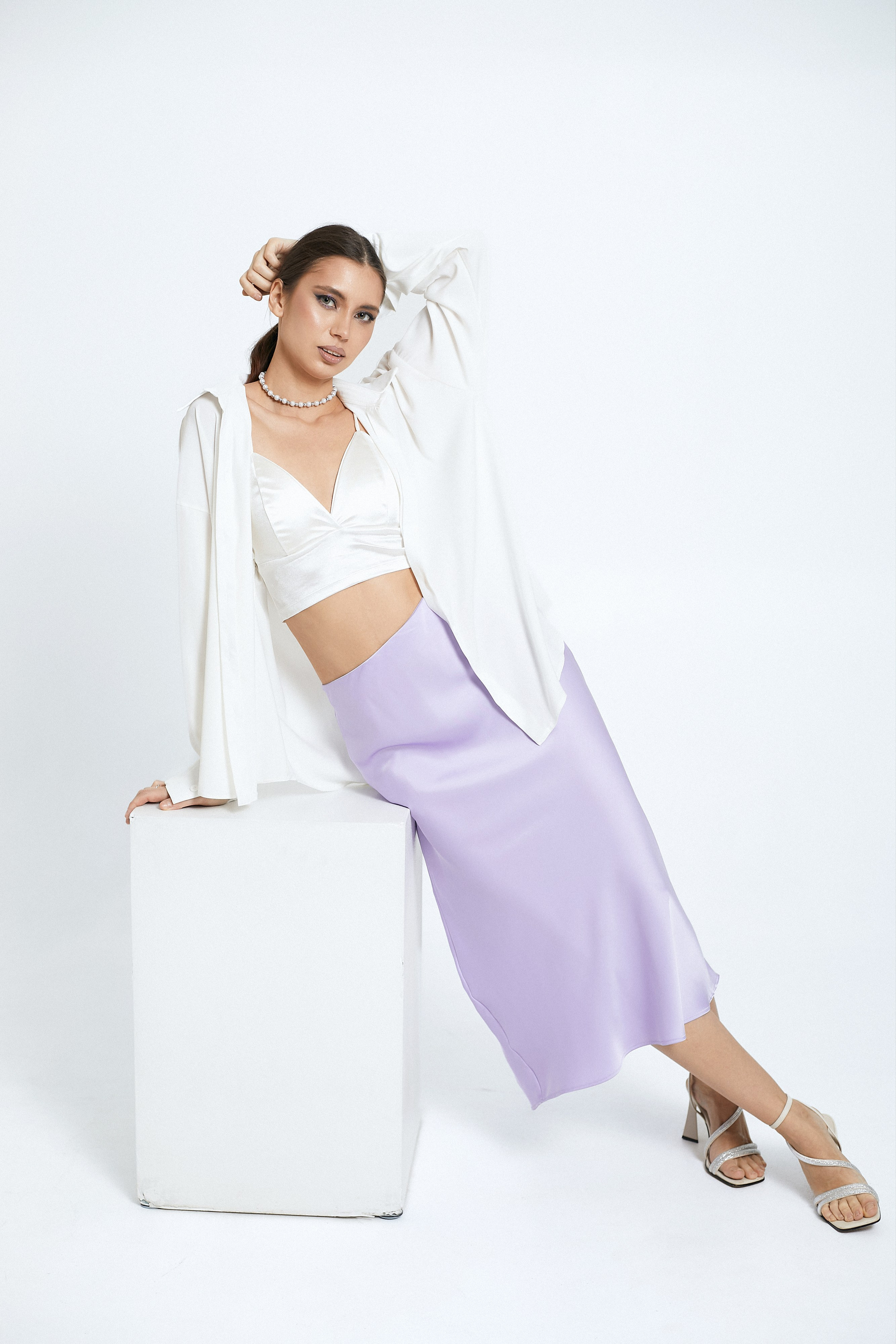 Красивая девушка в белом атласном топе, рубашке и сиреневой юбке с белым кубом