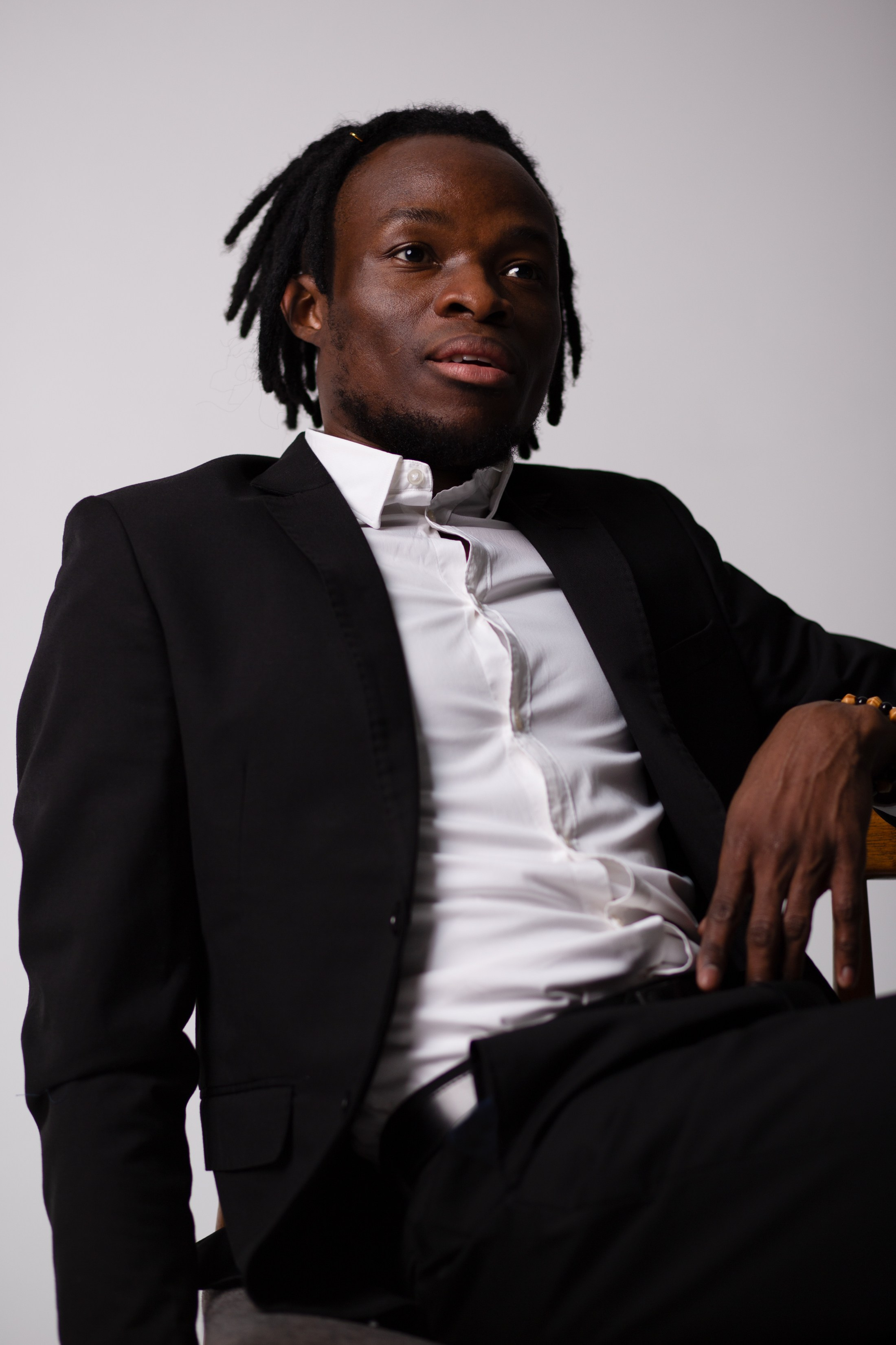 Чернокожий парень в черном костюме и белой рубашке сидит на стуле.