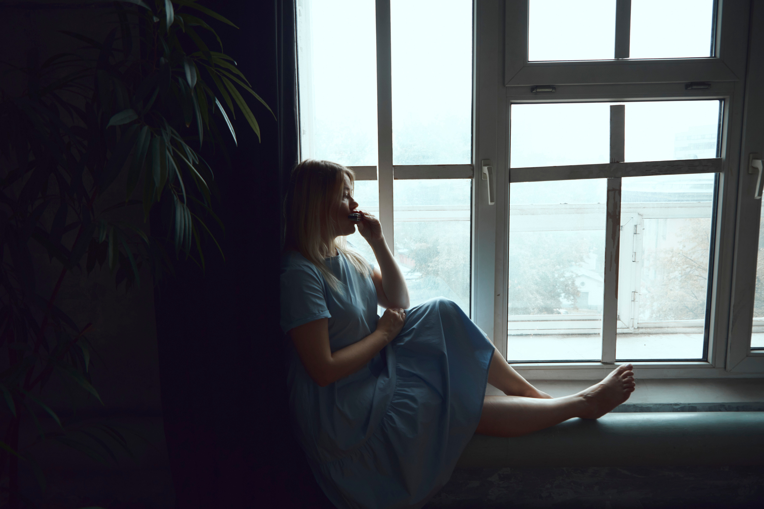 Девушка в синем платье сидит на подоконнике и играет на губной гармошке. На фоне окна хорошо виден её силуэт