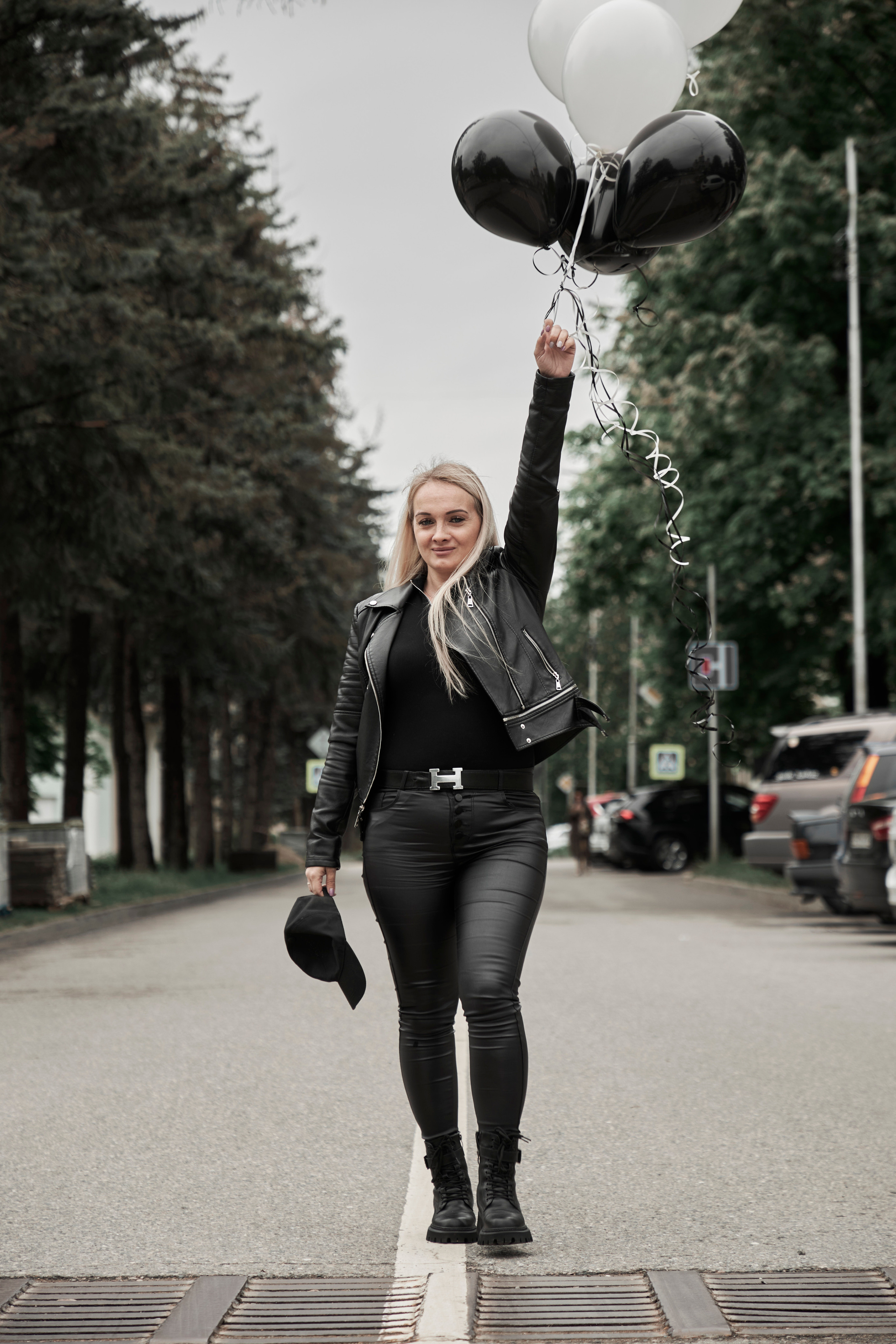 Красивая блондинка идёт по дороге и держит в руках черные  и белые воздушные гелиевые шары на вытянутой вверх руке