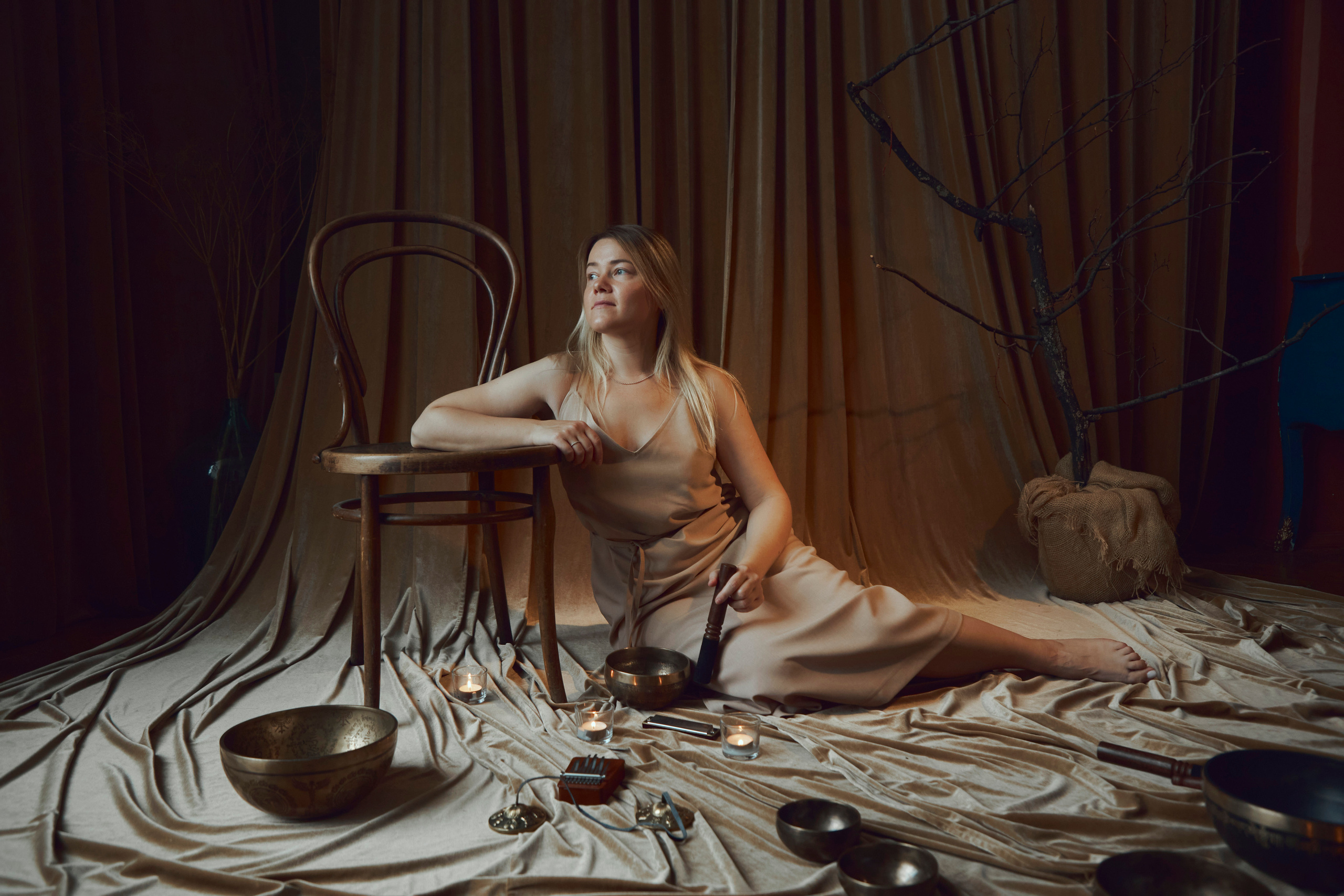 Молодая девушка сидит на полу опираясь рукой на стул в окружении атрибутов звукотерапевта