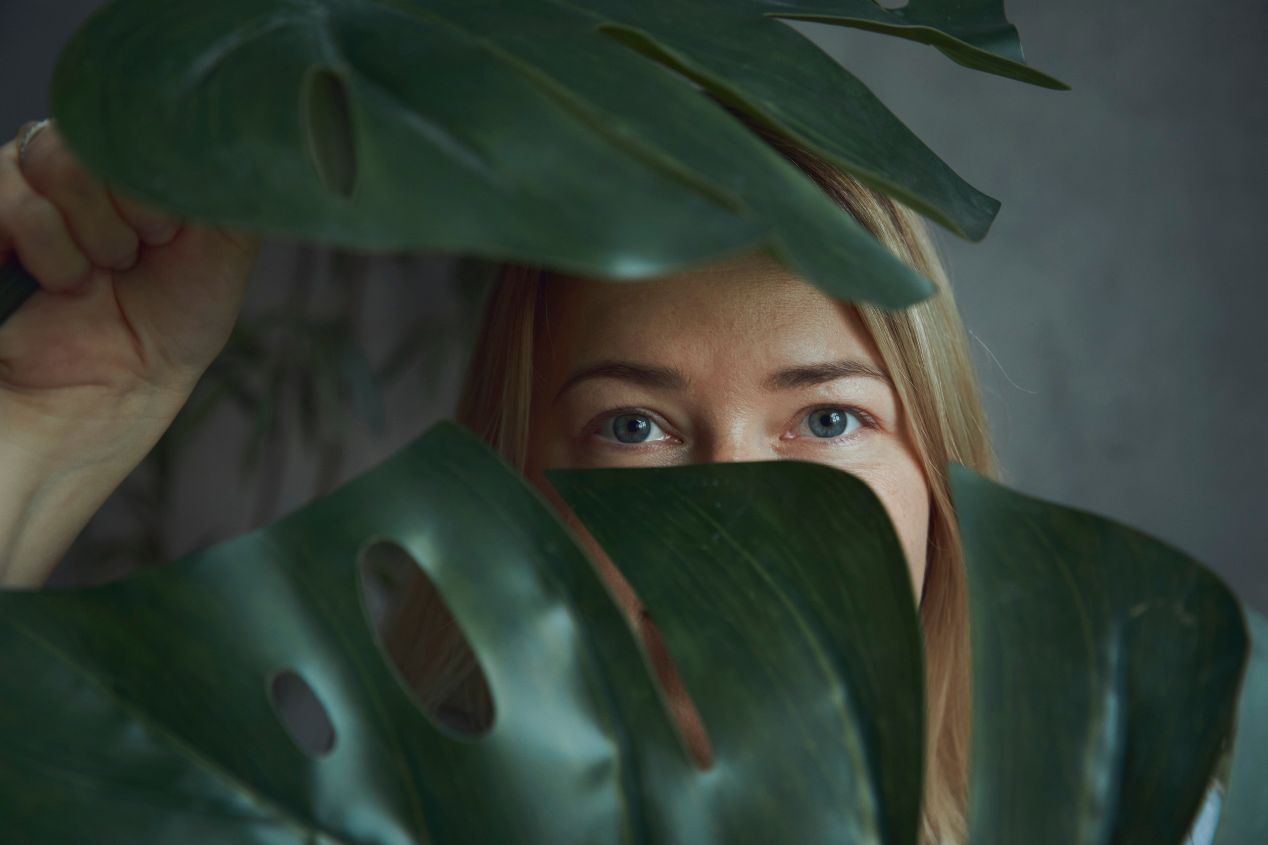 Девушка держит в руках листья монстерры так, что сквозь них видны только её глаза