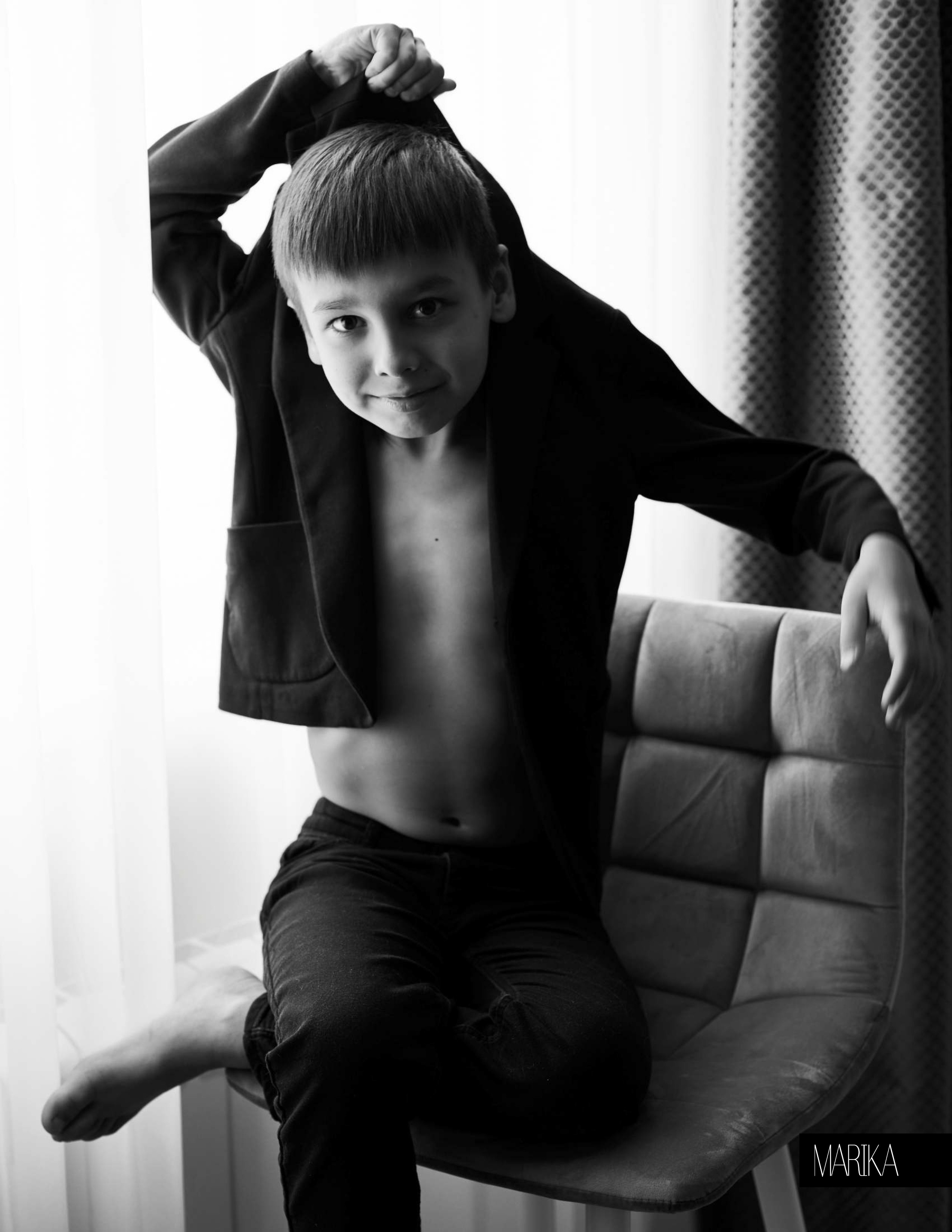 Портрет мальчика, сидящего на стуле около окна. Мальчик тянет себя за воротник пиджака вверх, как Барон Мюнхгаузен