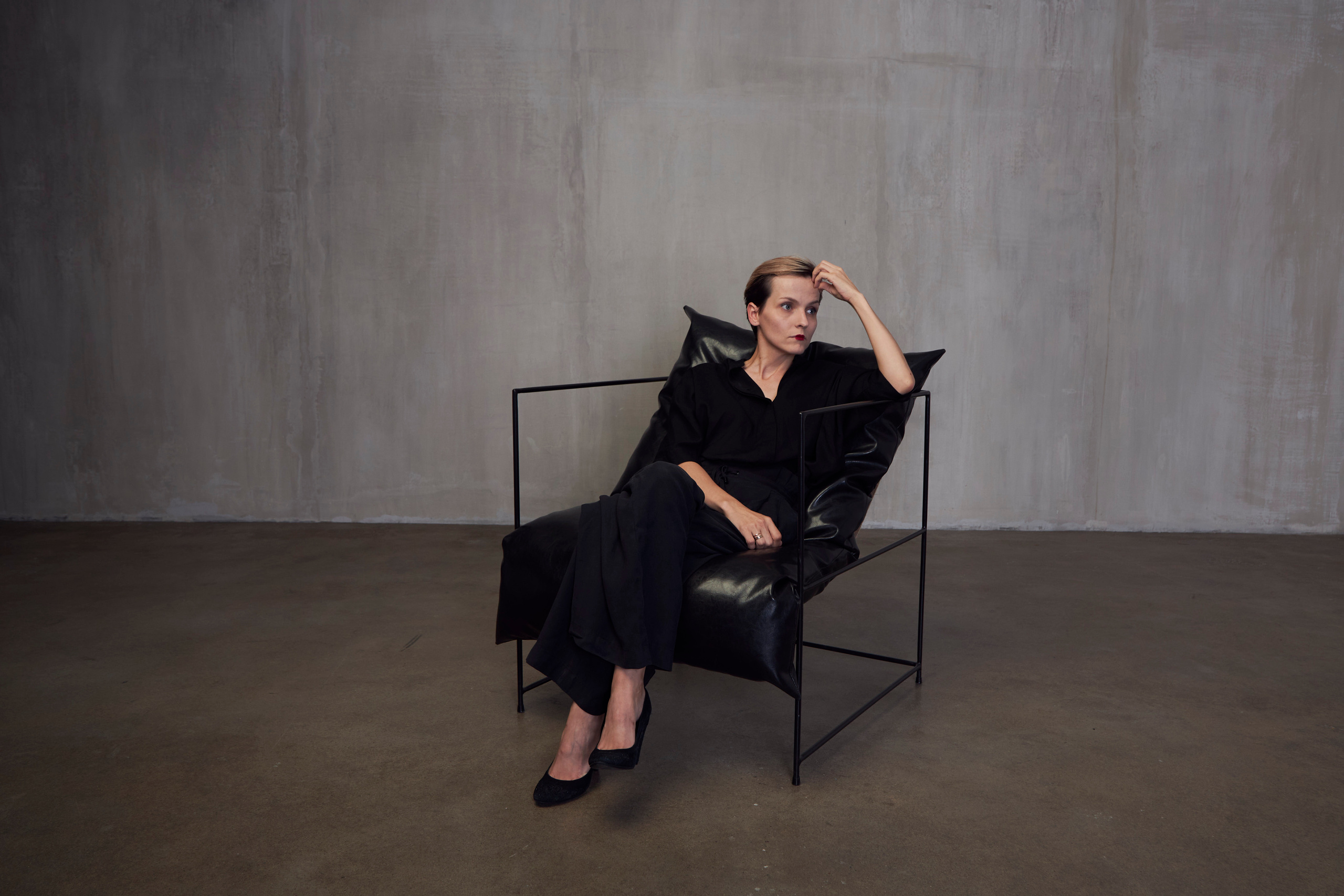 Девушка в черном костюме сидит в черном кожаном кресле на фоне серой стены в студии