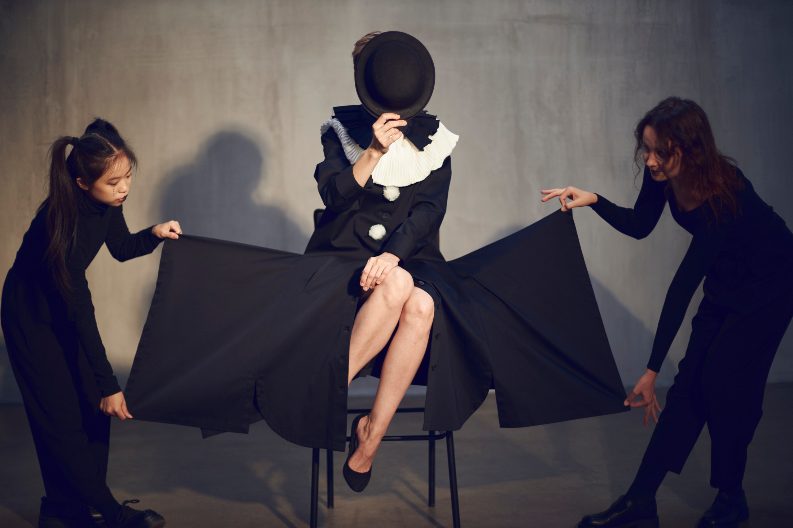 Девушка в черном платье сидит на стуле и прикрывает лицо шляпой, а её за полы платья держат  две девушки