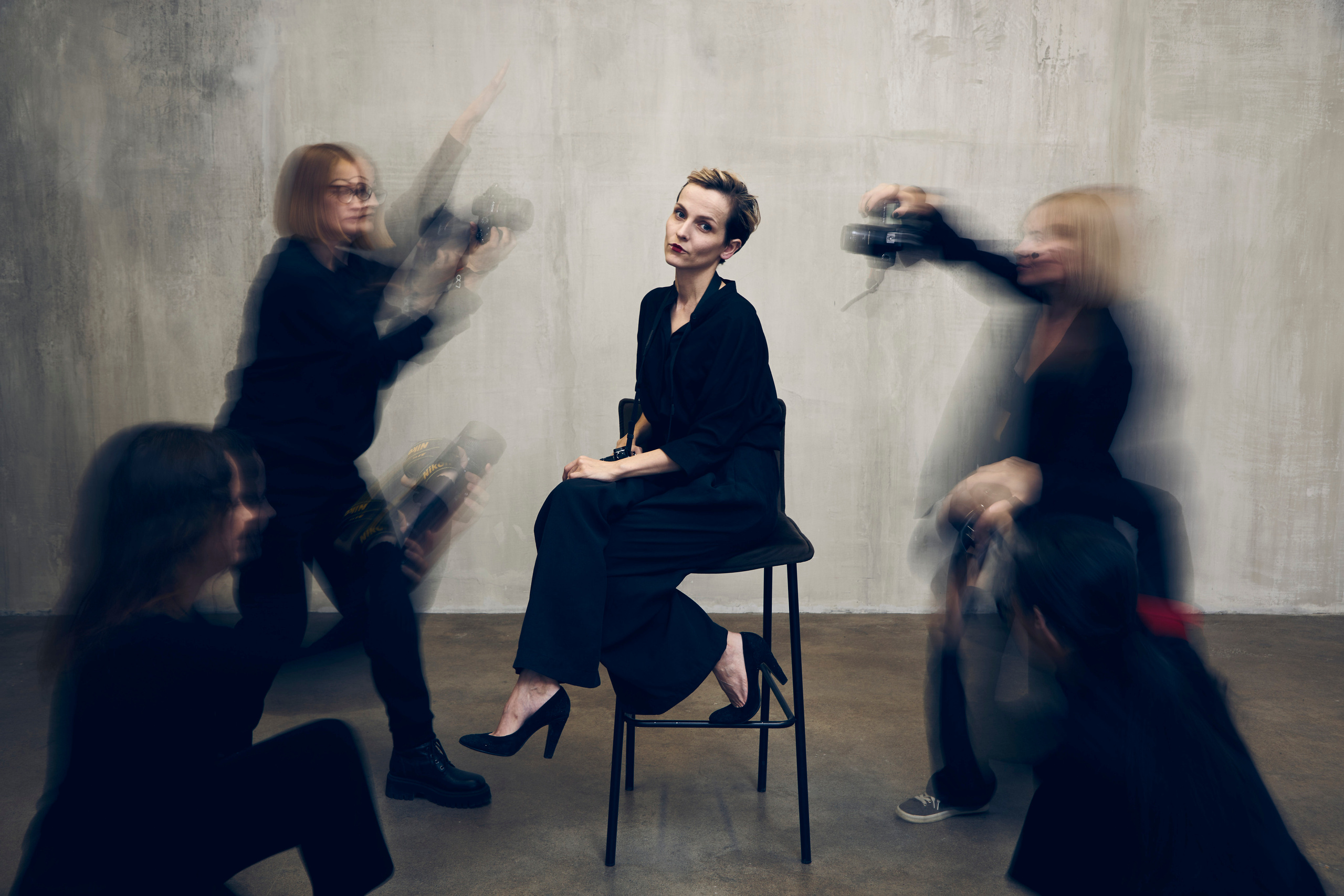 Девушка в черном костюме сидит на стуле и улыбается, а её окружают размытые силуэты фотографов 