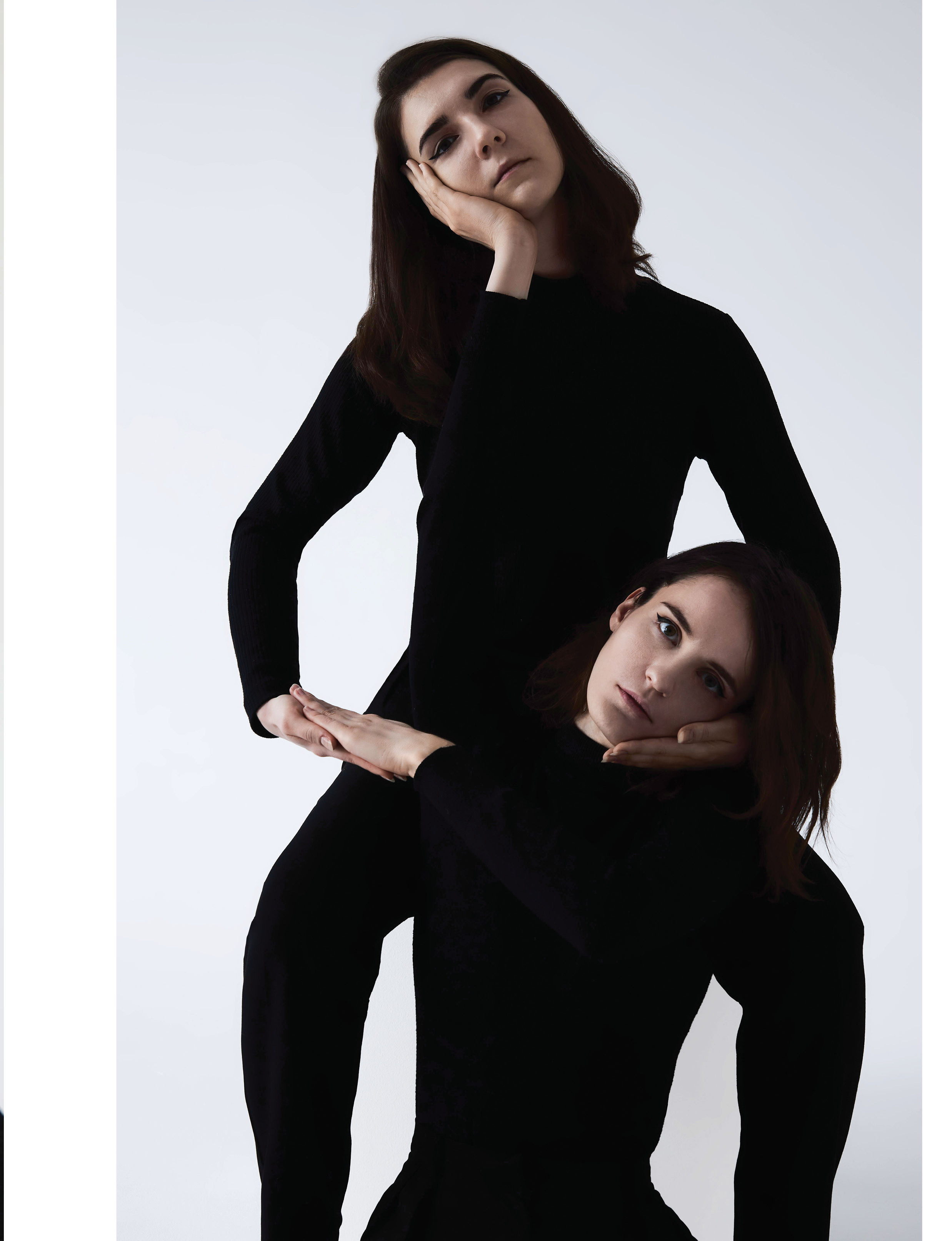 Две девушки в черных водолазках и черных брюках держат друг друга под подбородок