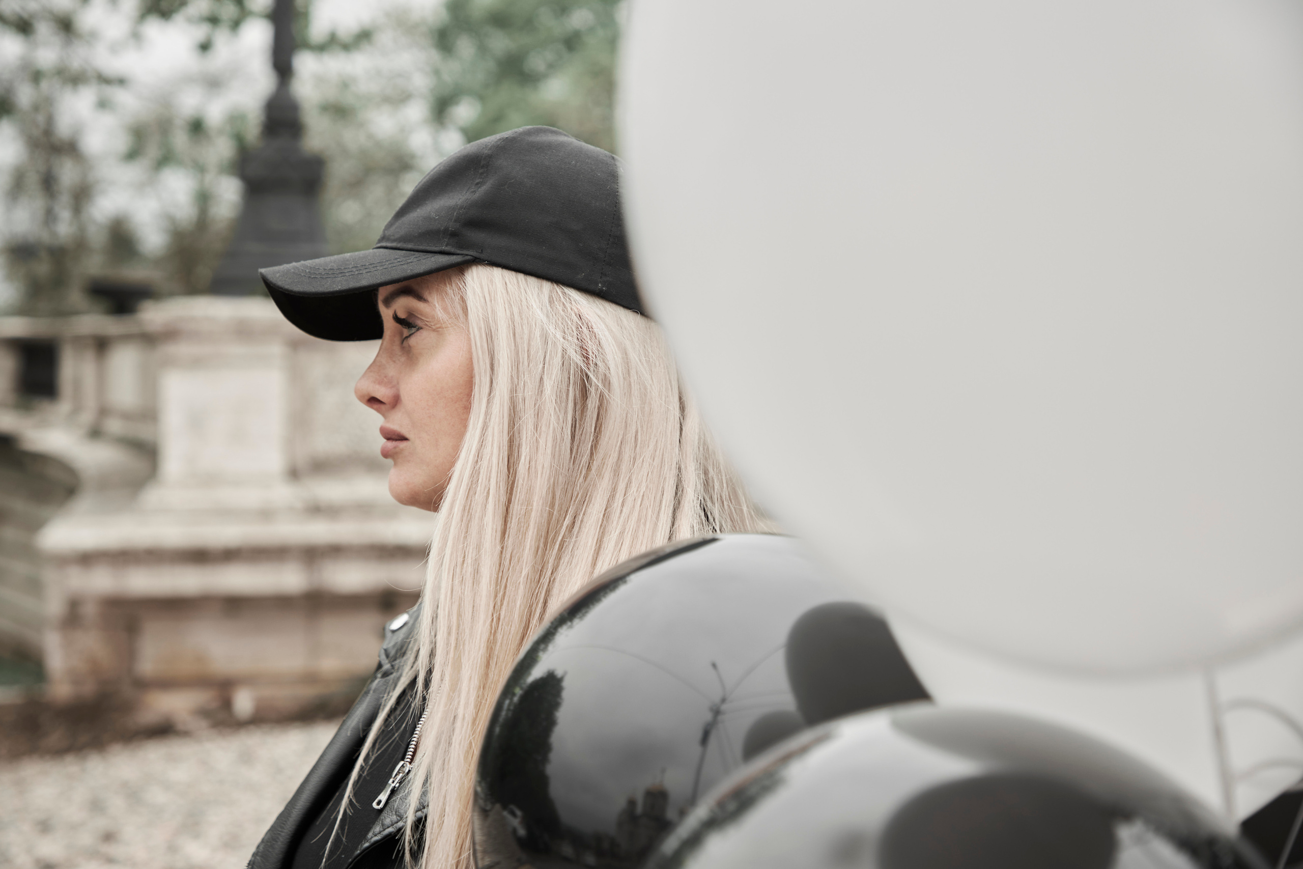 Девушка в кожаной кепке и с гелиевыми шариками стоит на дороге, портрет по плечи, в профиль