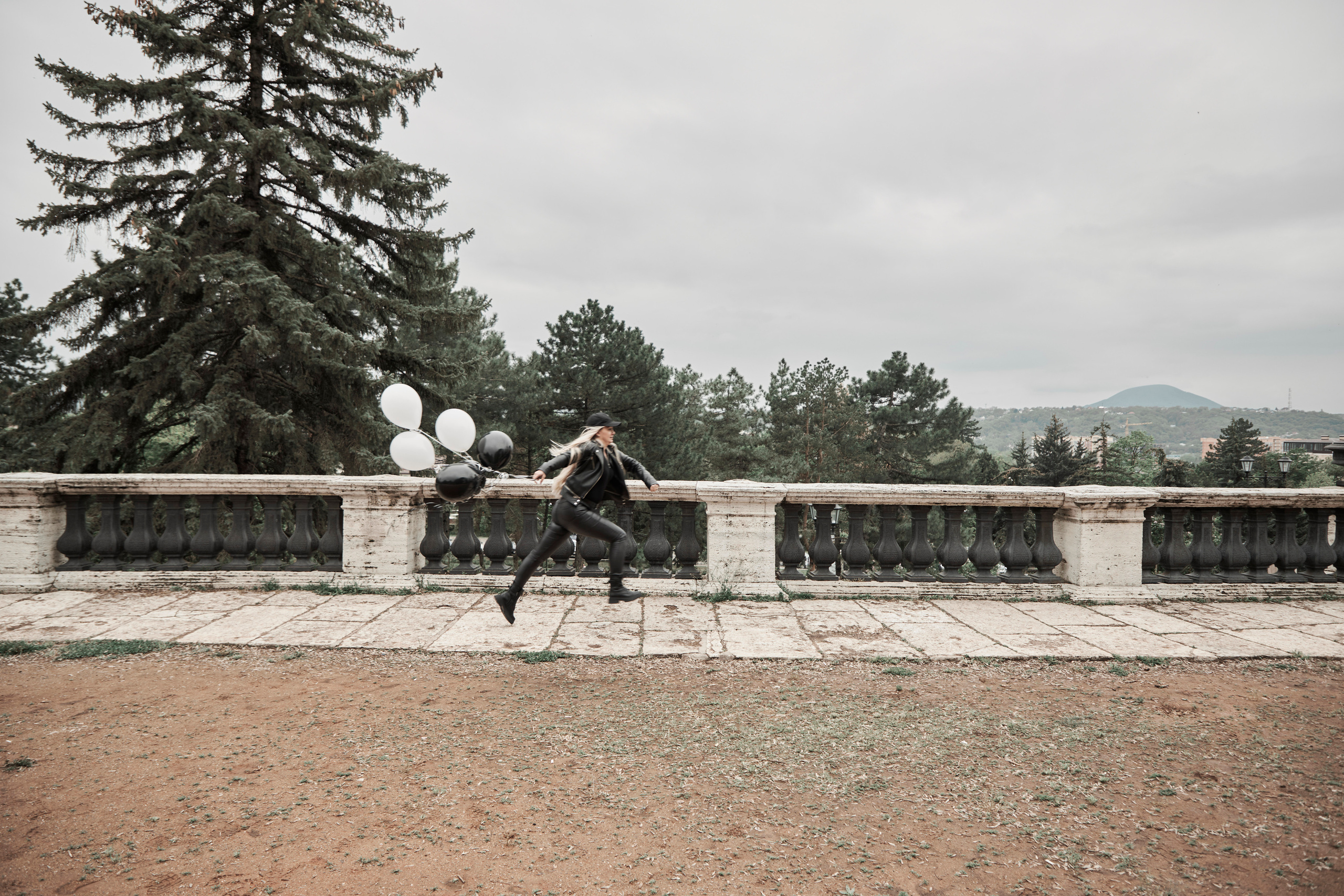 Общий план: девушка бежит по дорожке на фоне каменного парапета и держит в руках гелиевые шарики