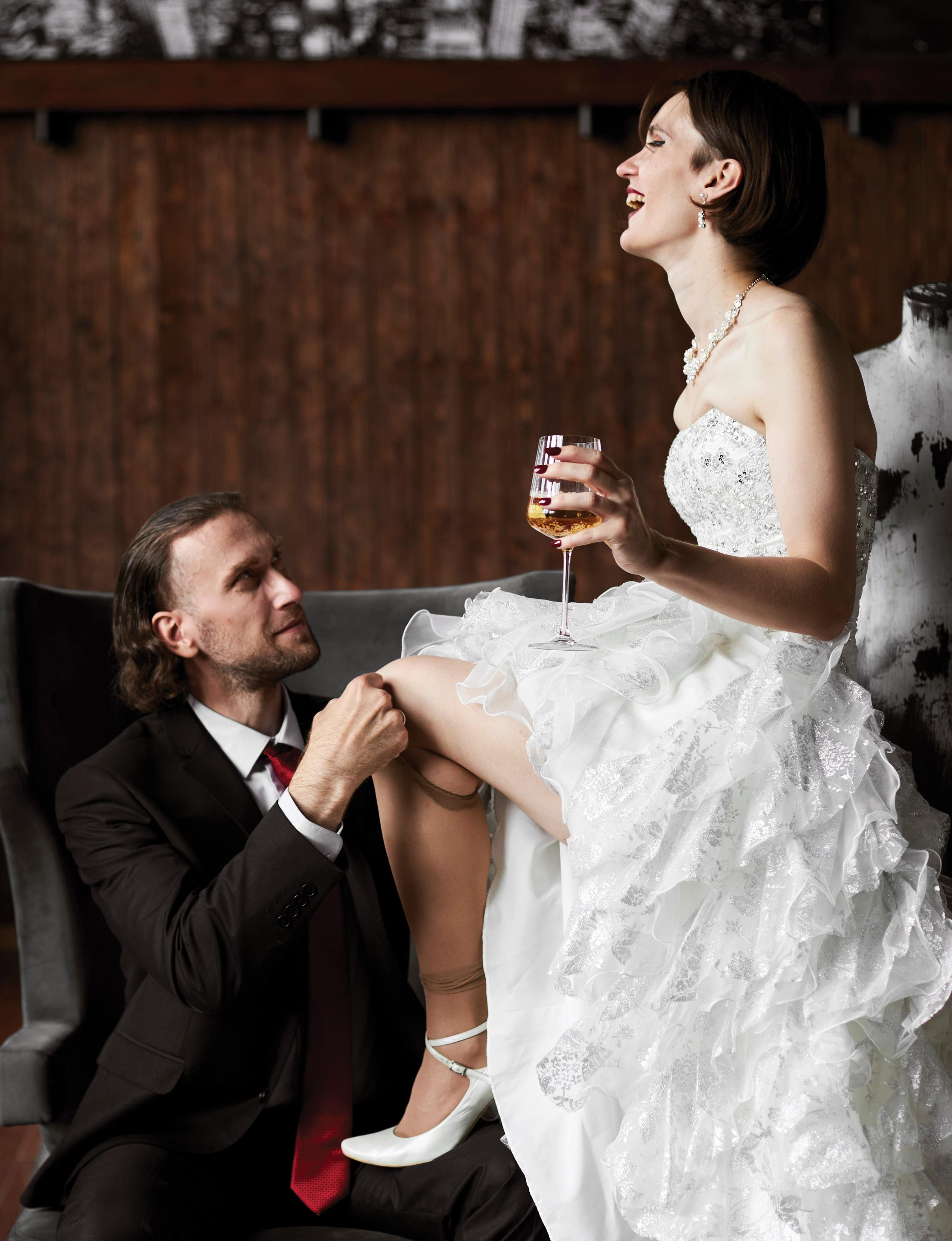 Влюбленная пара молодоженов на фотосессии в студии. Невеста пьёт шампанское, а жених снимает с неё чулки