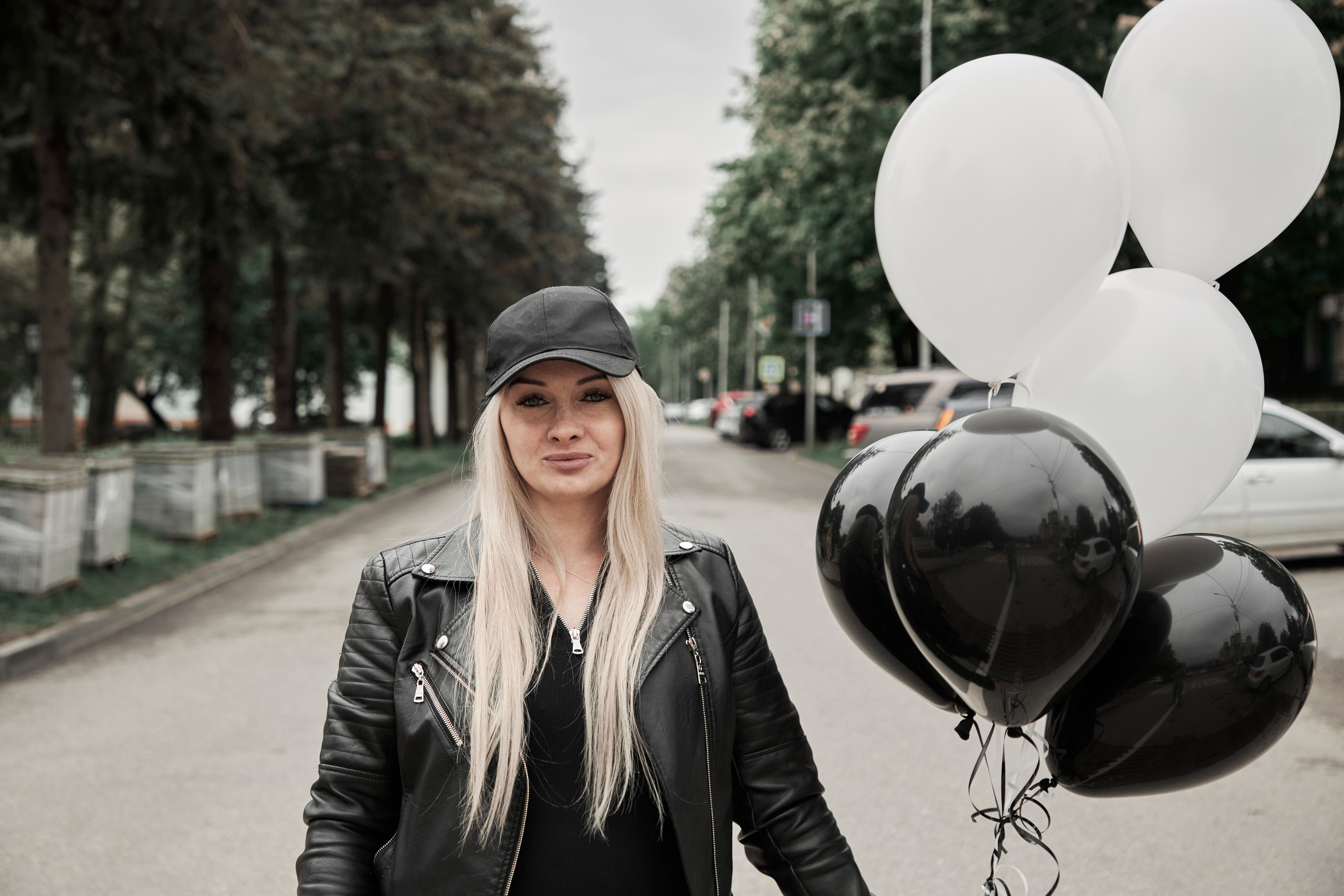 Девушка в кожаной кепке и с гелиевыми шариками стоит на дороге, портрет по пояс