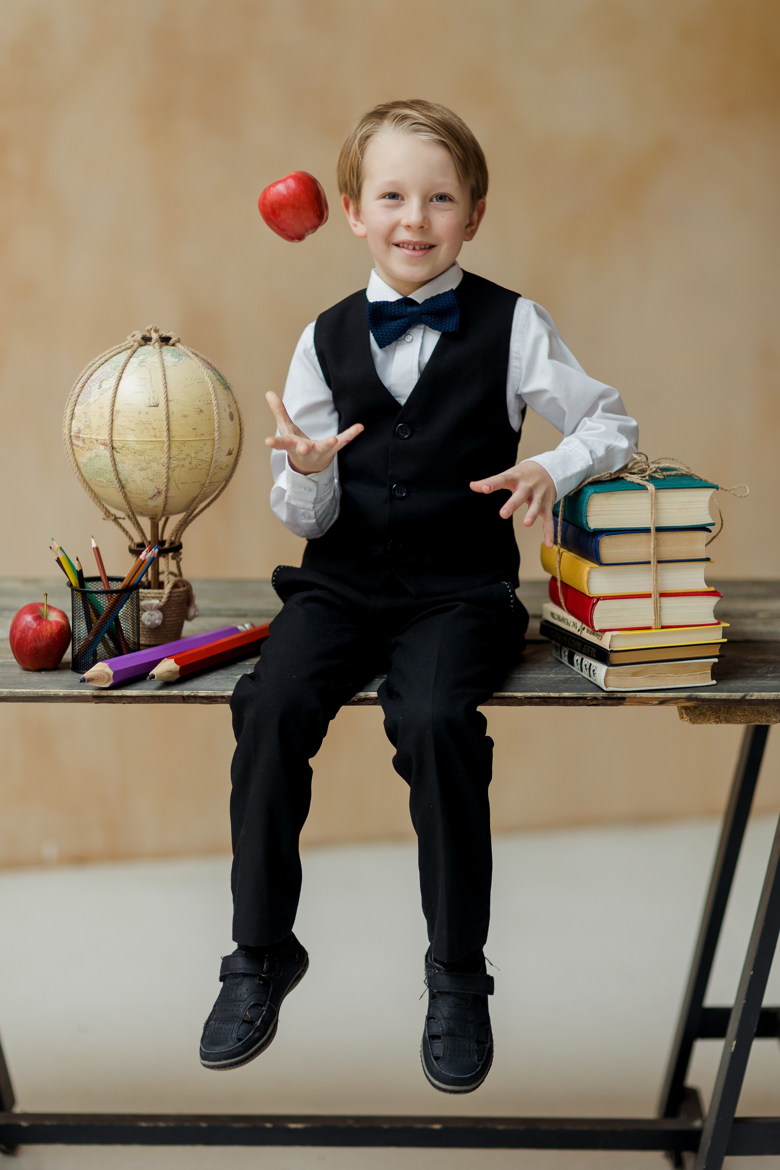 Мальчик сидит на столе с книжками и подкидывает яблоко