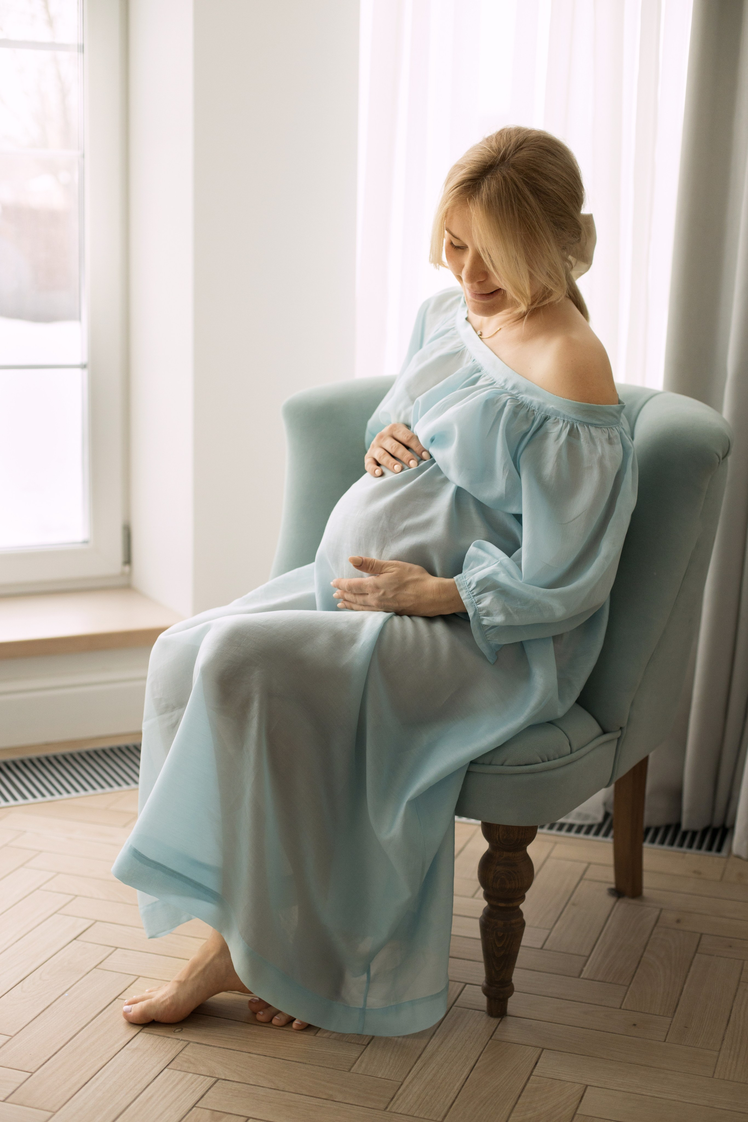 Беременная женщина в голубом платье сидит в кресле у окна и поглаживает живот