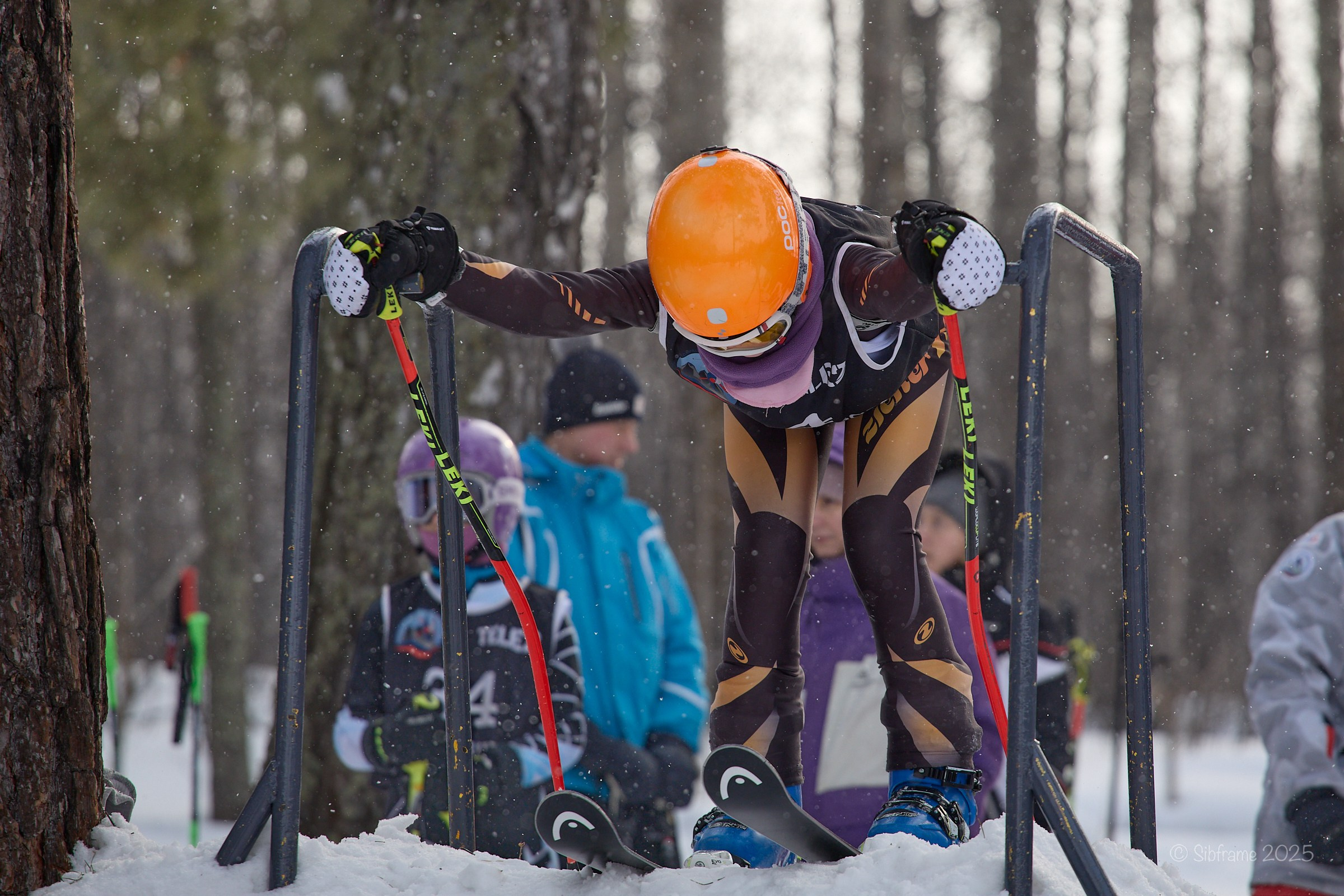 Юный участник ски-кросса в оранжевом шлеме и обтягивающем костюме на стартовых воротах