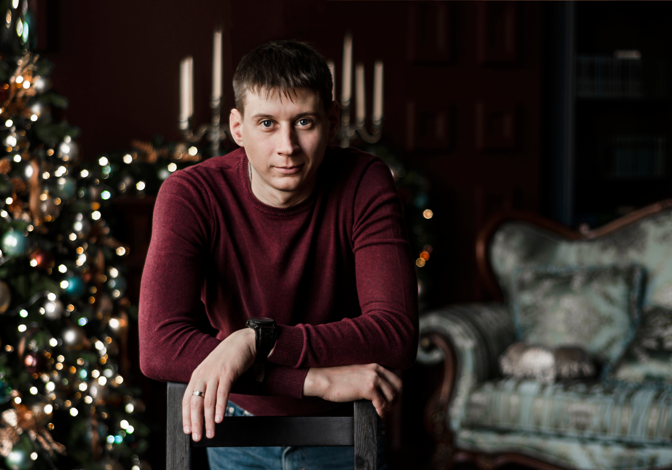 Молодой человек на фоне елки оперся на спинку стула.