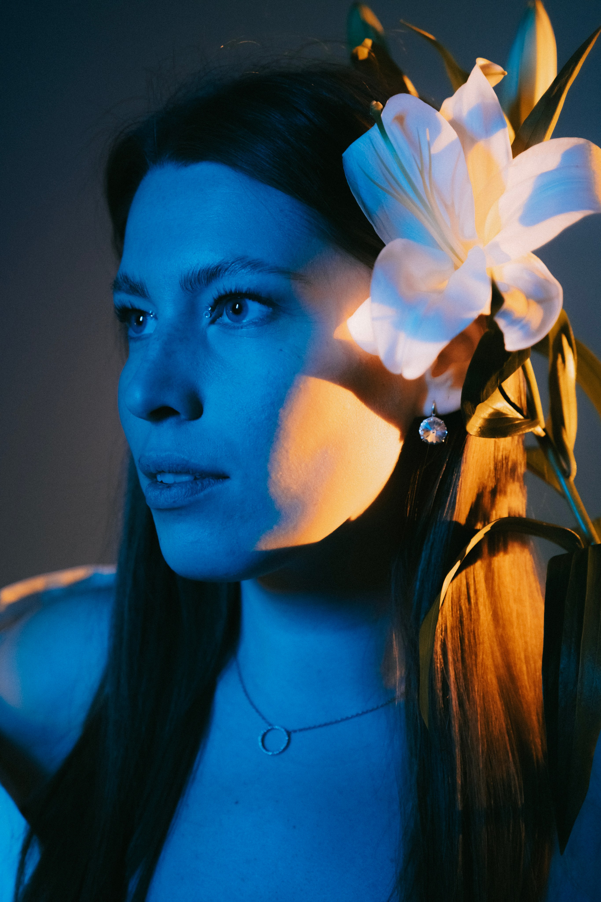 Портрет молодой женщины с белой лилией в волосах, в художественном освещении синим и тёплым светом, создающим контрастные тени на лице и цветке.