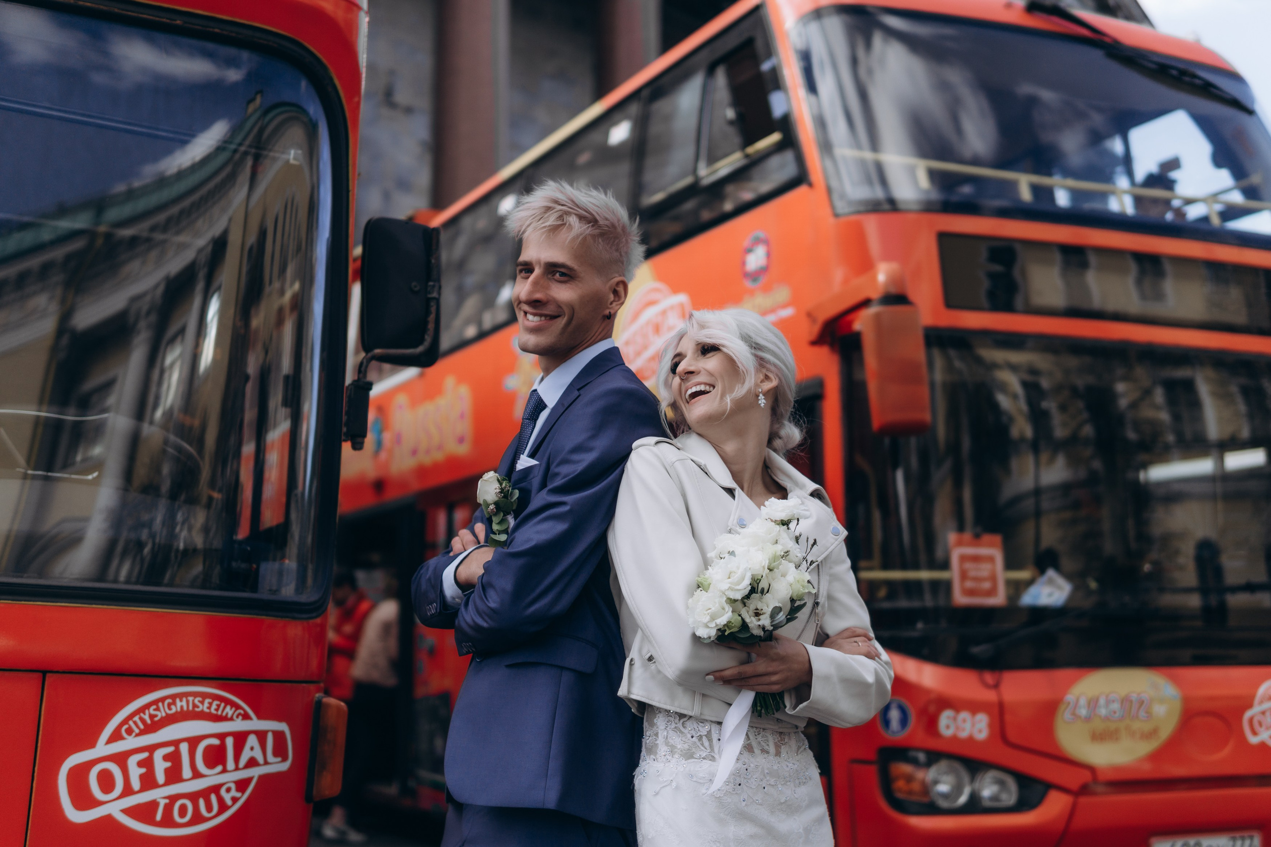 жених блондин в синем костюме и невеста блондинка стоят спиной к спине на фоне красных туристических автобусов