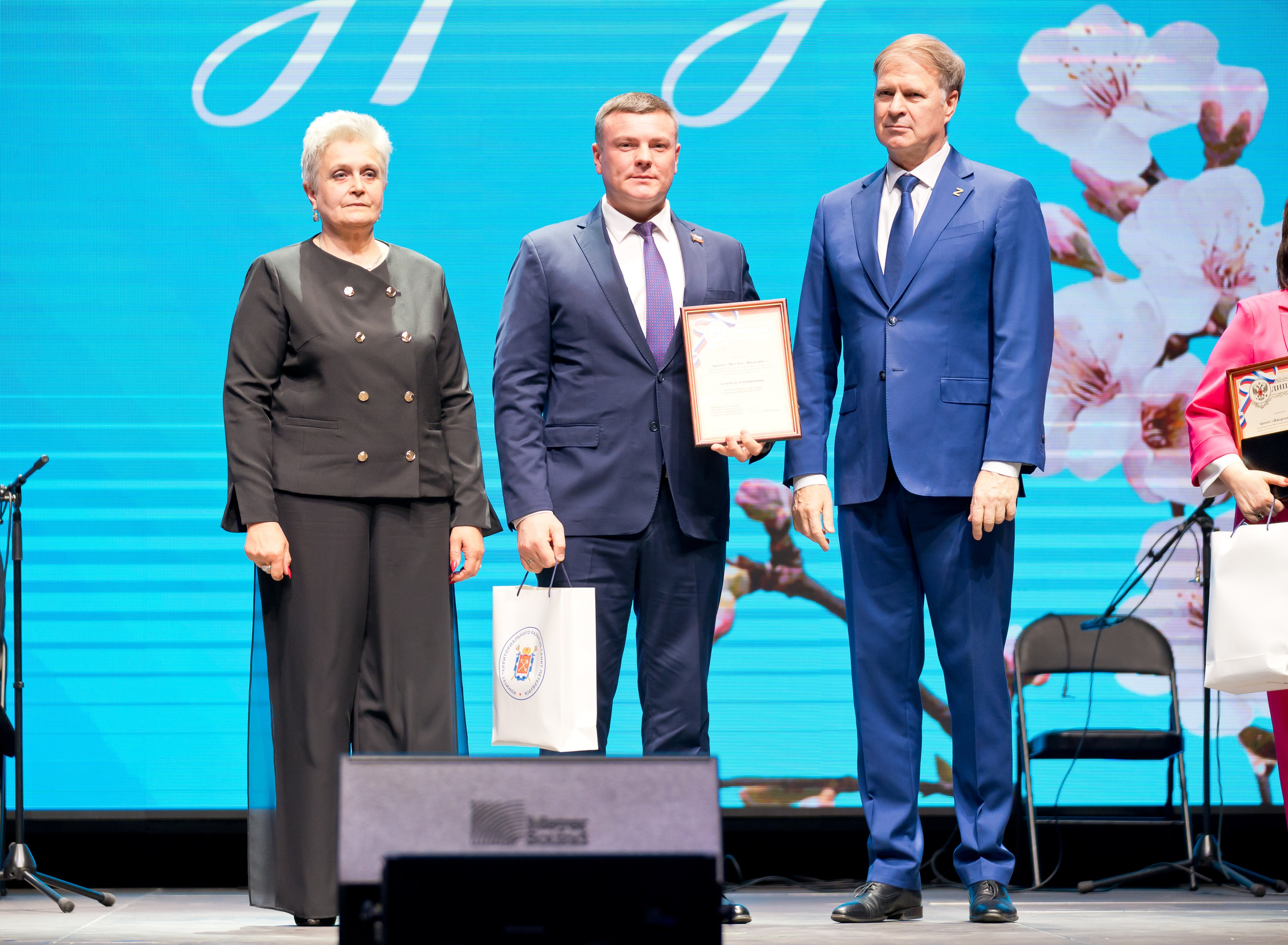 Награждение на сцене: женщина в чёрном костюме, лауреат и официальное лицо с грамотой. День местного самоуправления Санкт-Петербург | Фото: Евгений Мальцев.