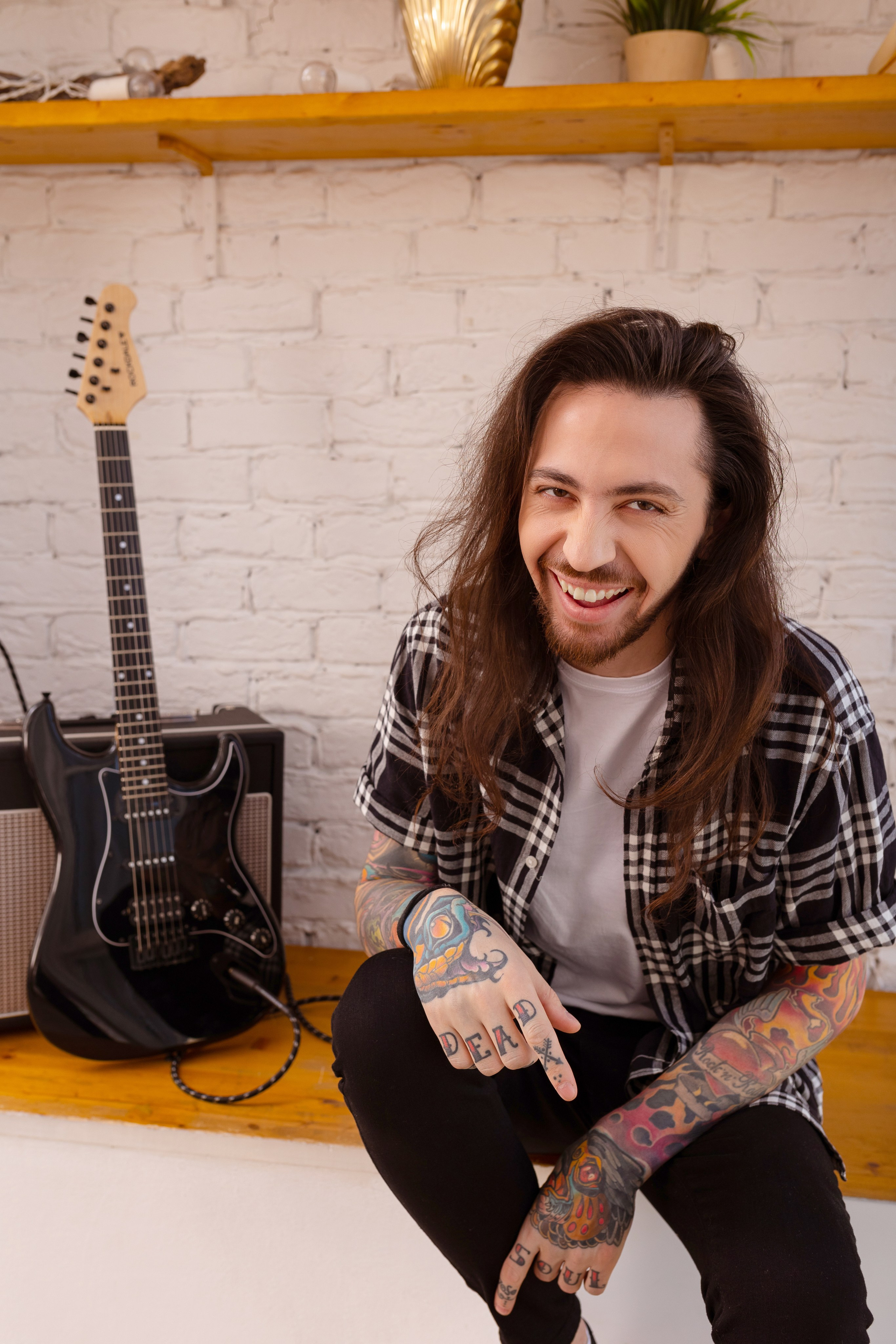 Молодой человек с длинными волосами сидит рядом с черной электрогитарой.