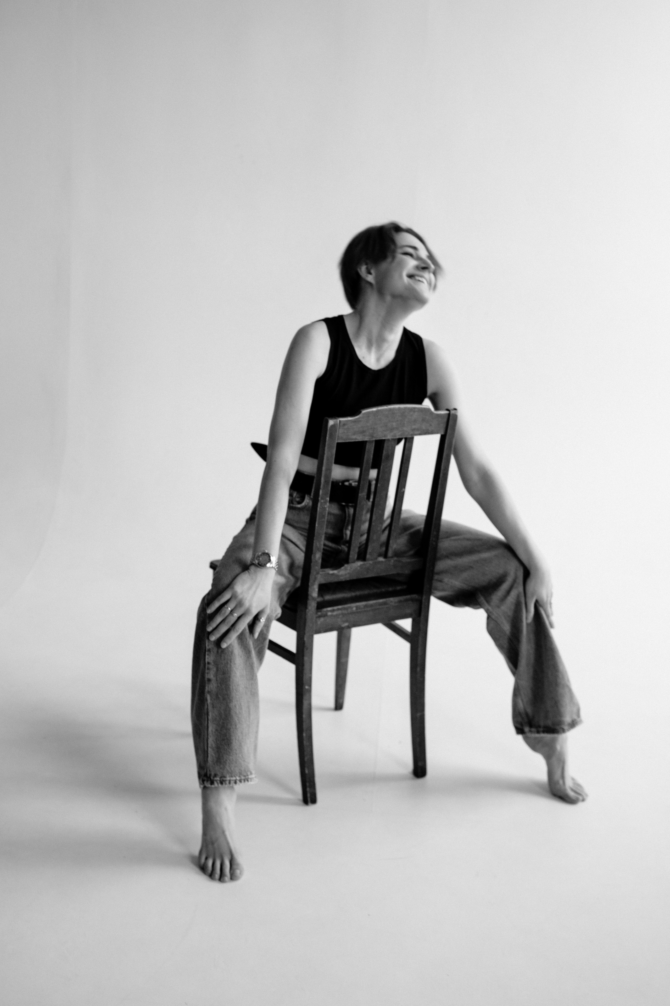 Чёрно-белое фото женщины, сидящей верхом на деревянном стуле в студии; она смеётся и наклоняется вперёд, упершись руками в спинку стула.