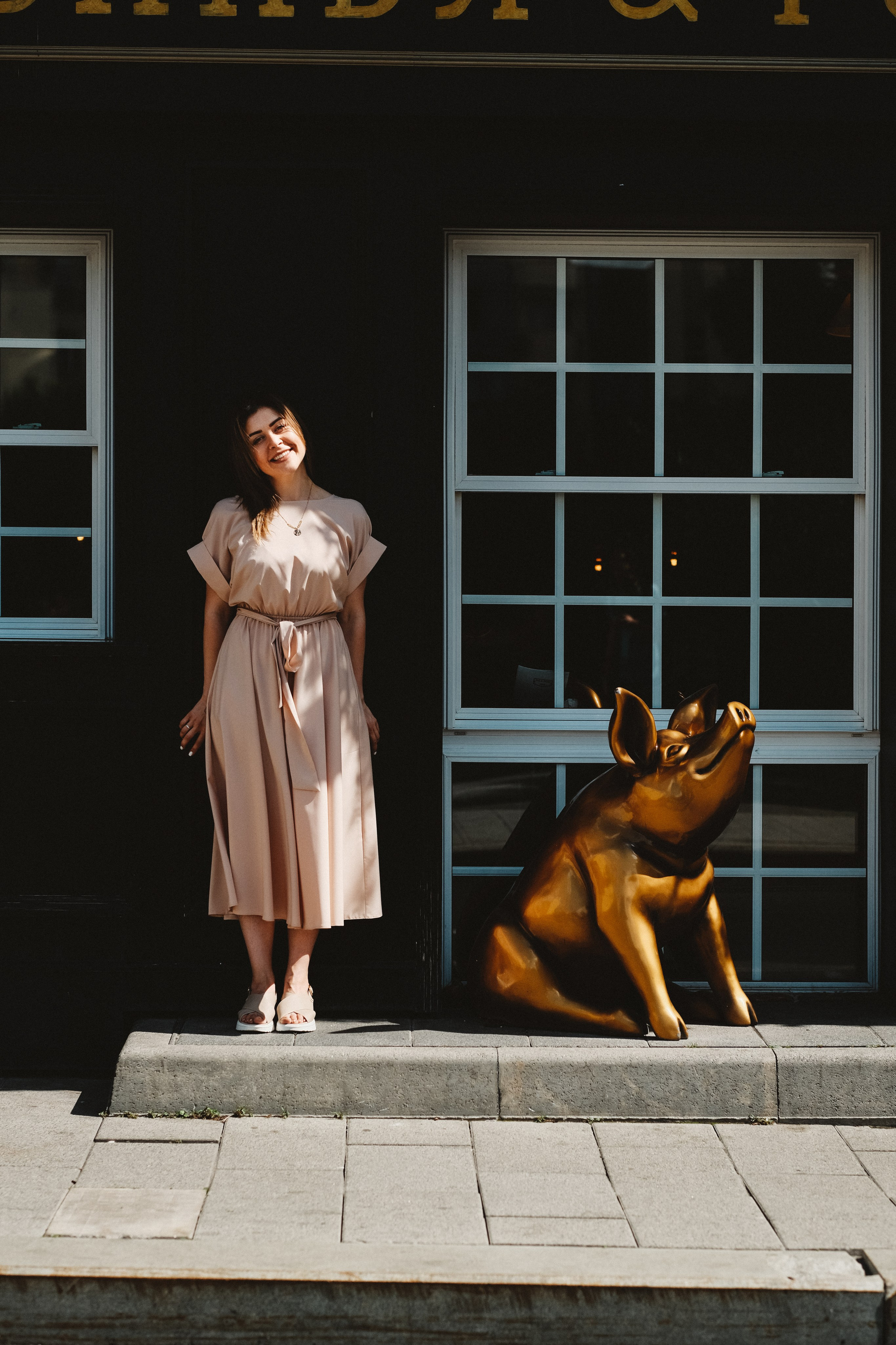 Девушка в бежевом платье на фоне старого черного дома, рядом с золотой свиньёй. Ресторан Свинья и Роза.