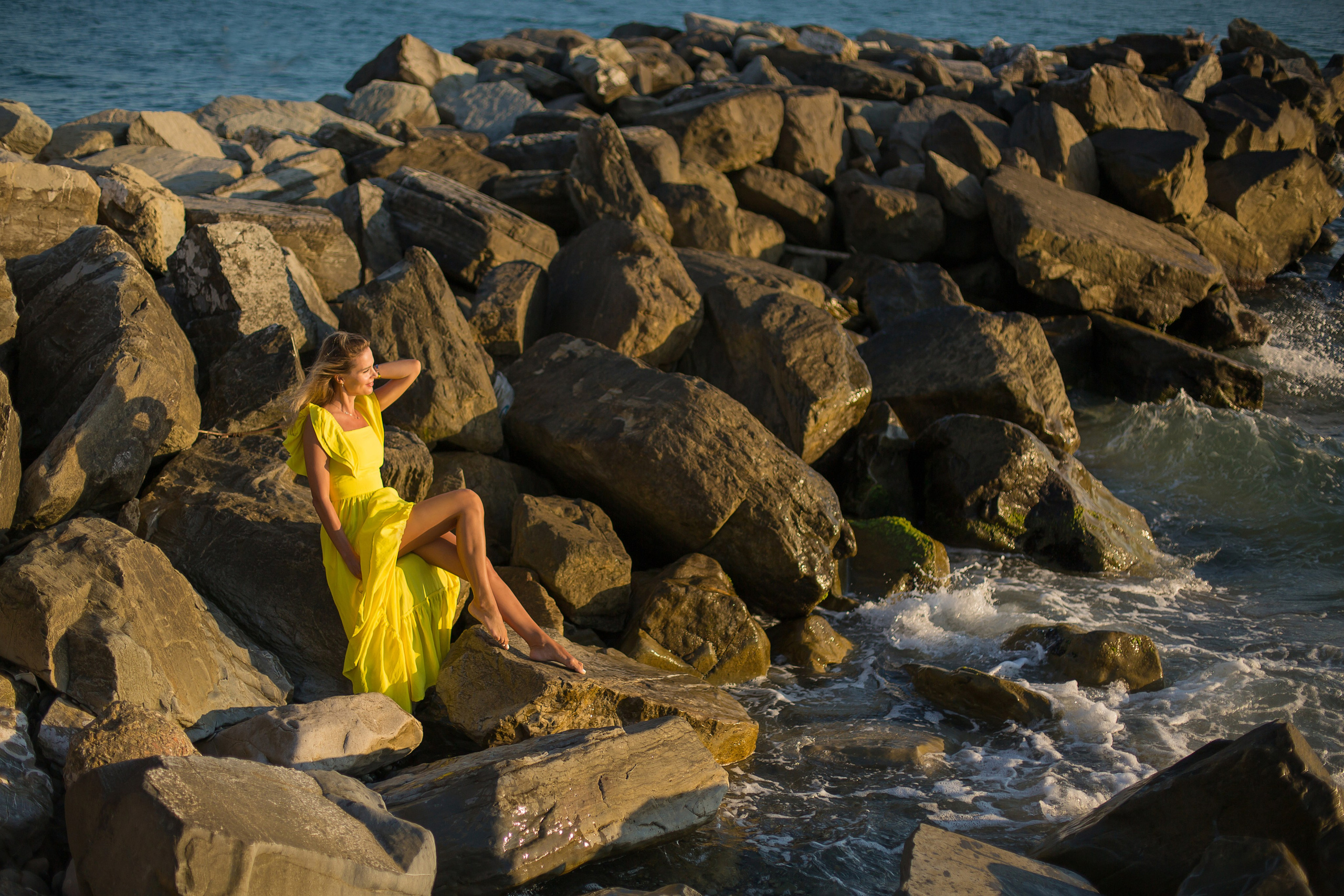 девушка в желтом платье сидит на больших камнях у моря