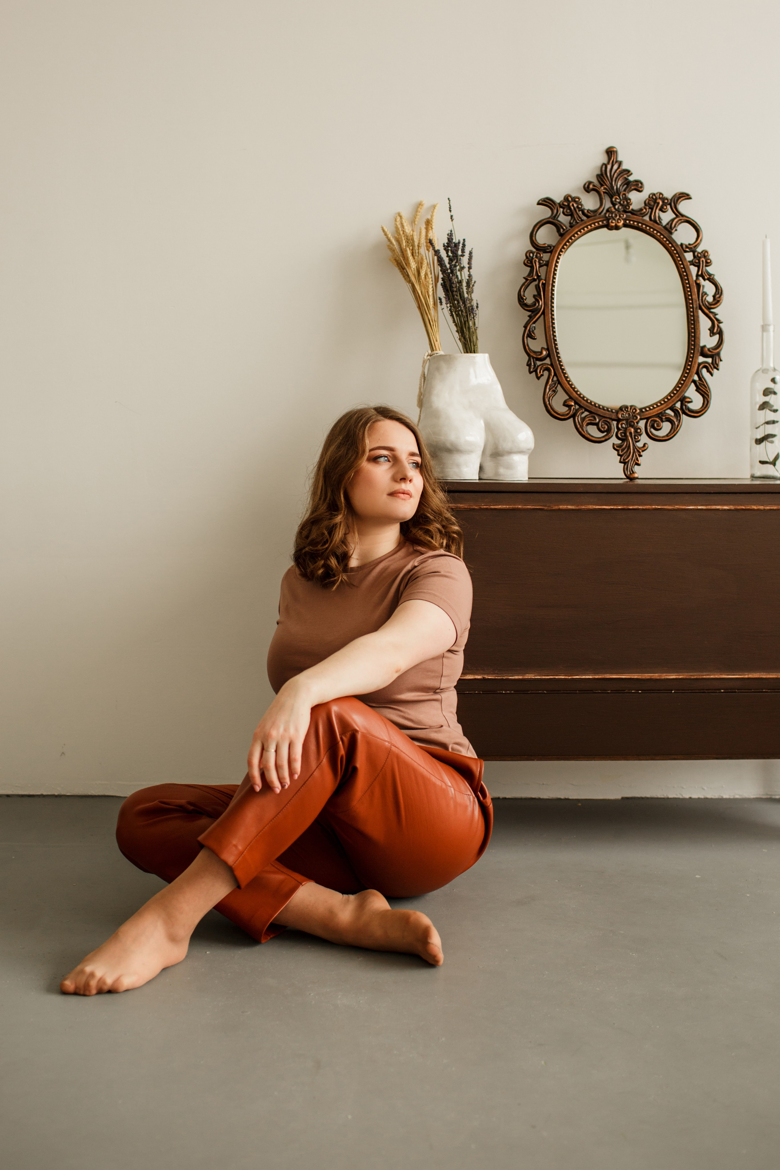 Цветной портрет девушки, сидящей на полу со скрещёнными ногами, в коричневом топе и кожаных брюках цвета терракоты, на фоне винтажного зеркала и пианино.