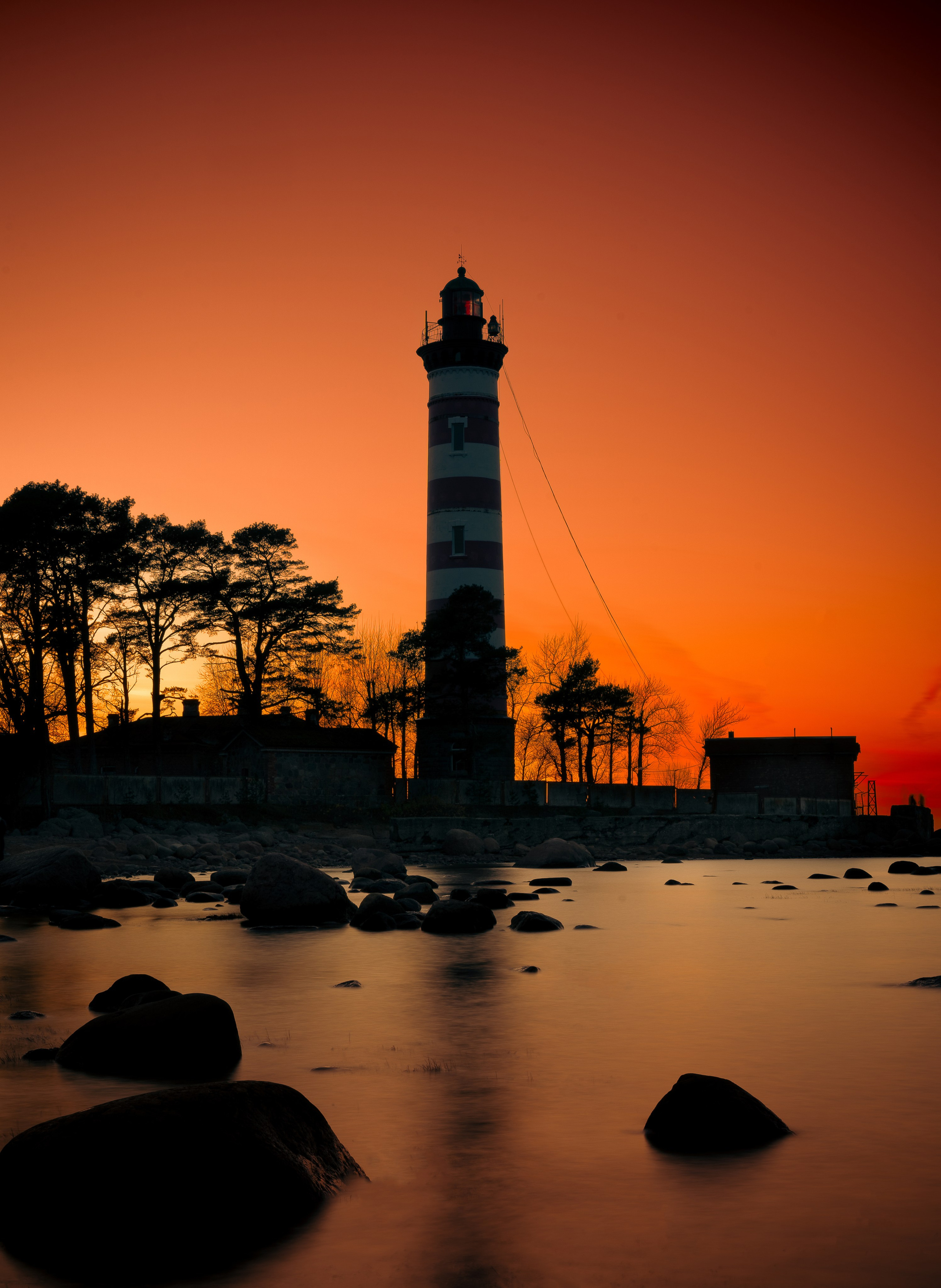 Шепелевский маяк. Полосатый красный маяк силуэт на закате оранжевое небо Финский залив, сосны, камни, длинная выдержка, отражение воды фото Евгений Мальцев.