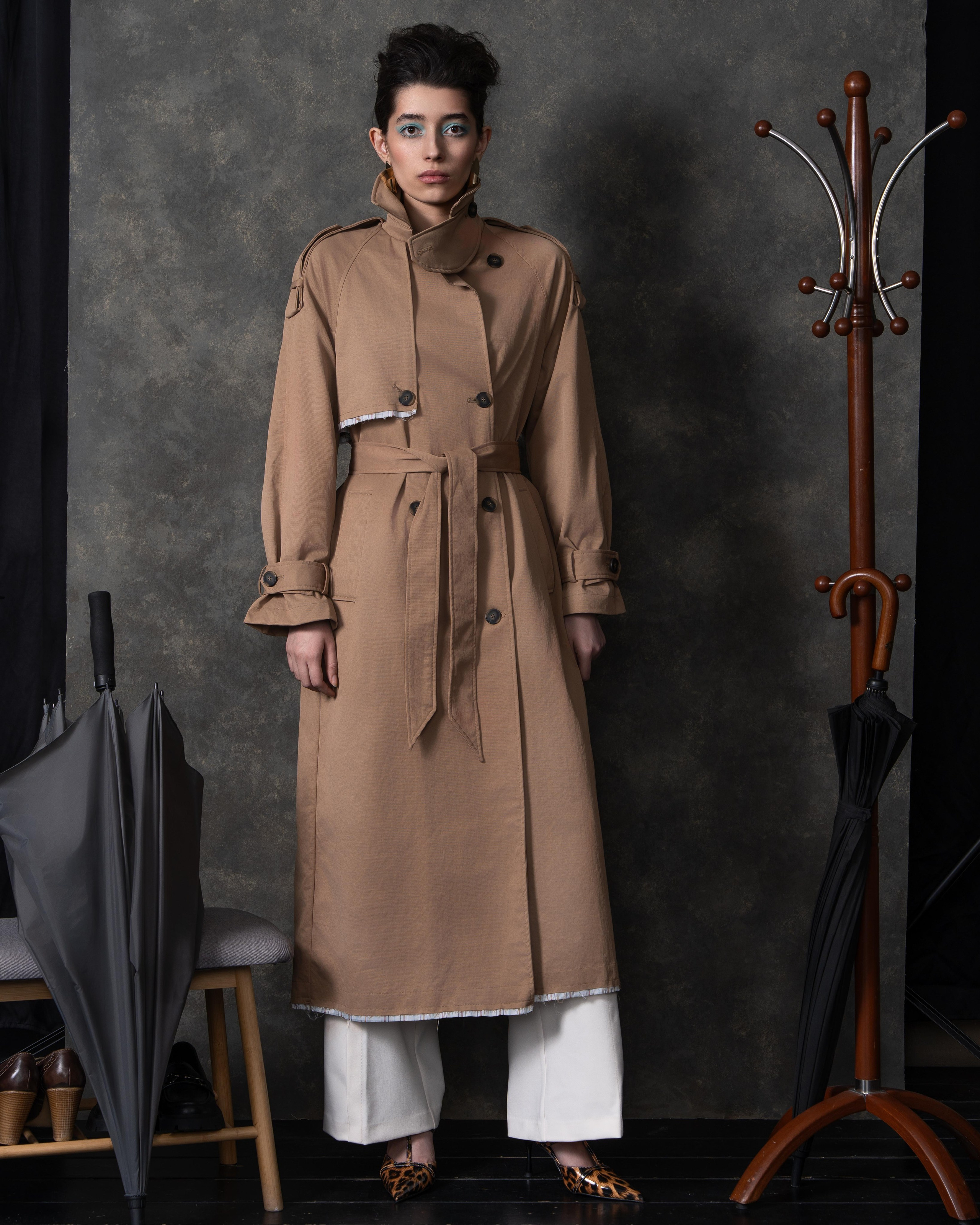 На фотографии изображена женщина в бежевом пальто с меховым воротником. Она стоит на фоне серой стены. На ней также белые брюки и туфли с леопардовым принтом. В одной руке она держит черный зонт, а в другой — коричневую шляпу. Рядом с ней стоит вешалка для одежды с несколькими зонтами.