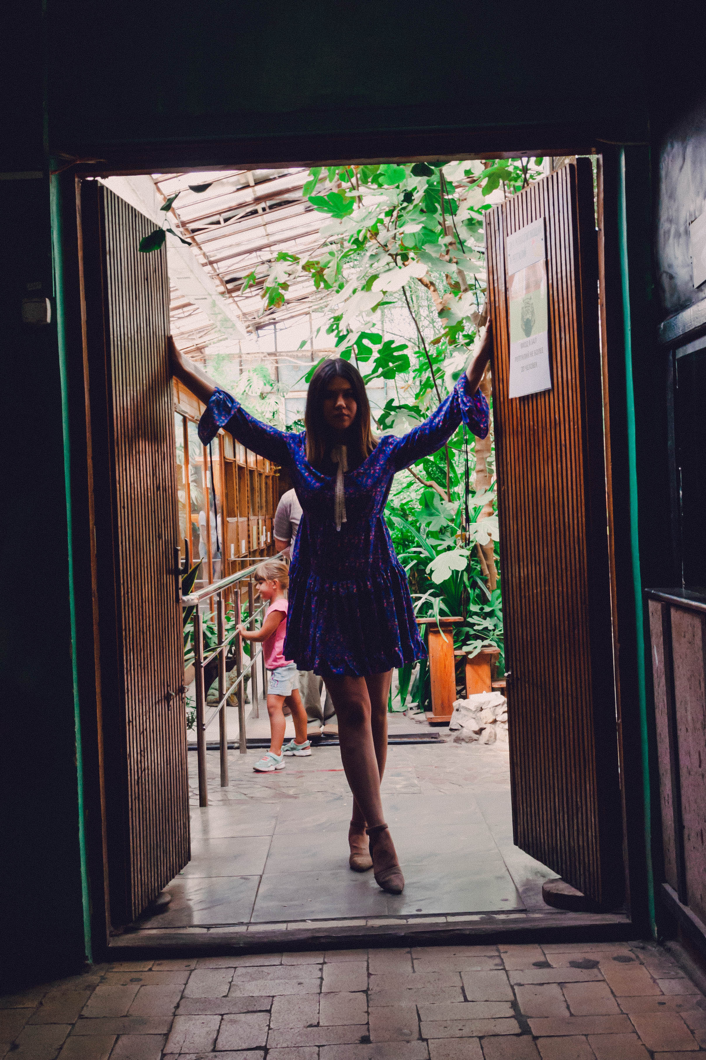 Женский силуэт в дверном проеме. Женщина в платье стоит в дверях, за ее спиной много зеленых растений.