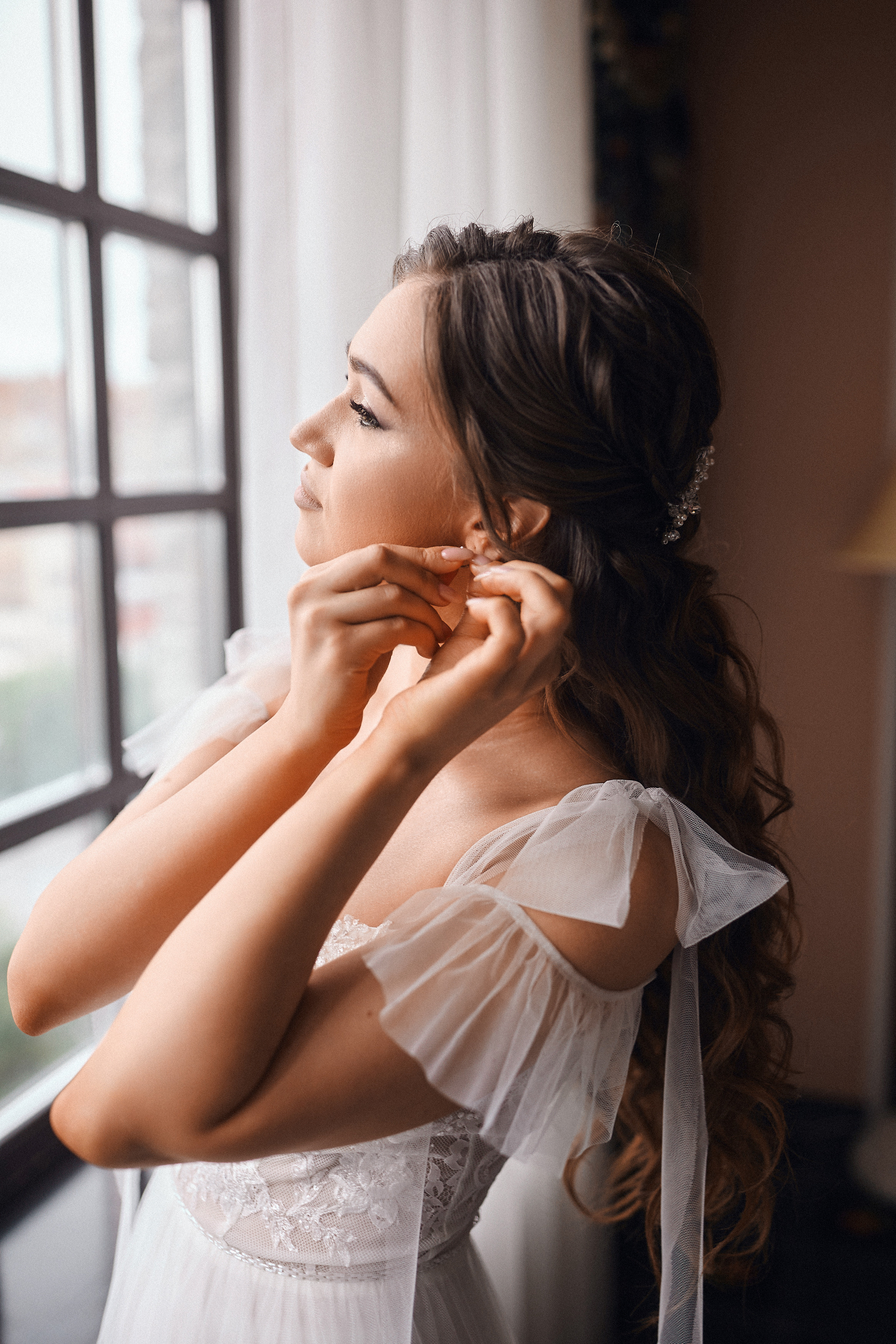 Красивая невеста у окна отеля надевает серьги