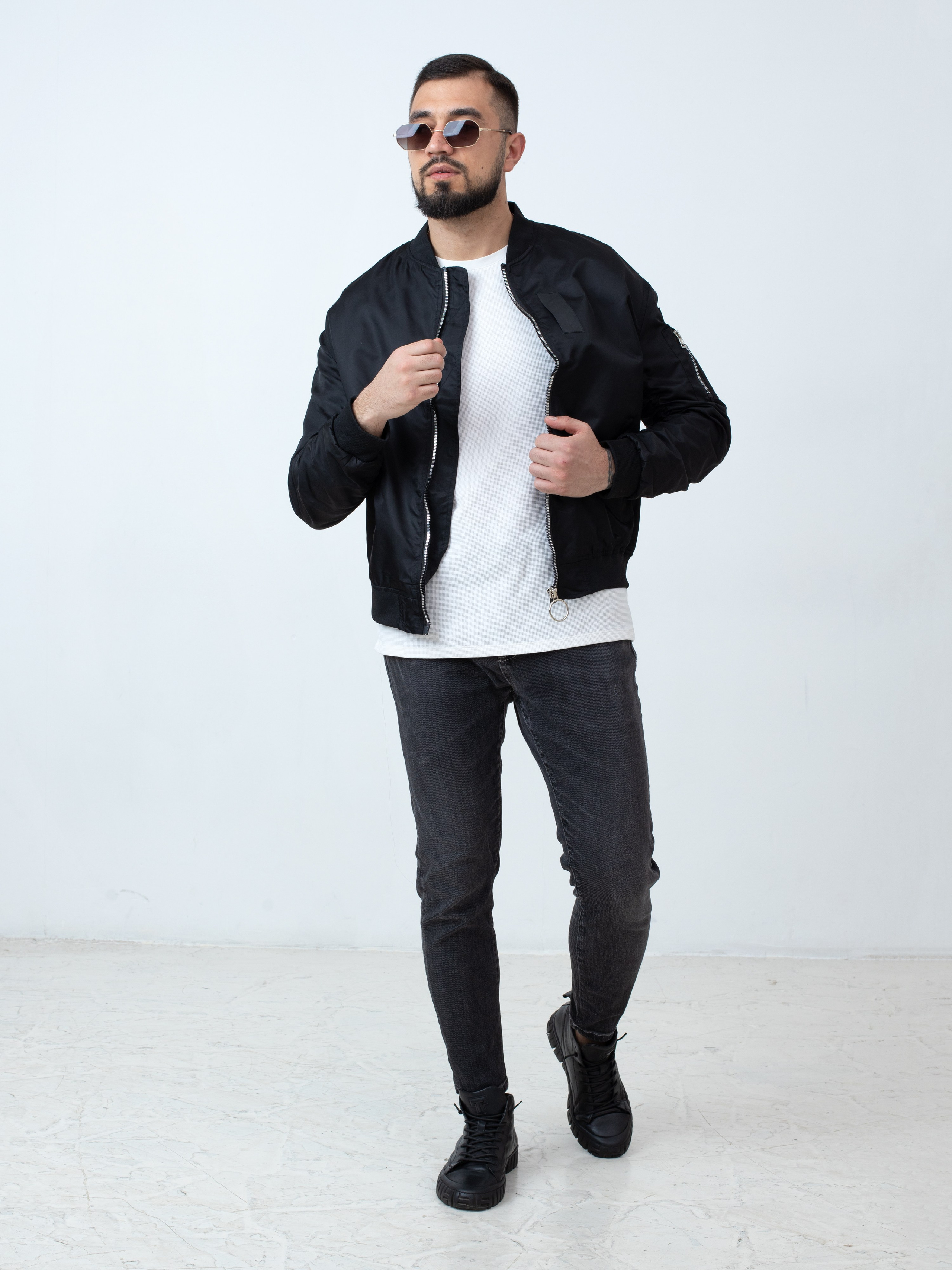 Мужчина брюнет с бородой в черной куртке бомбере, черных джинсах и белой футболке
