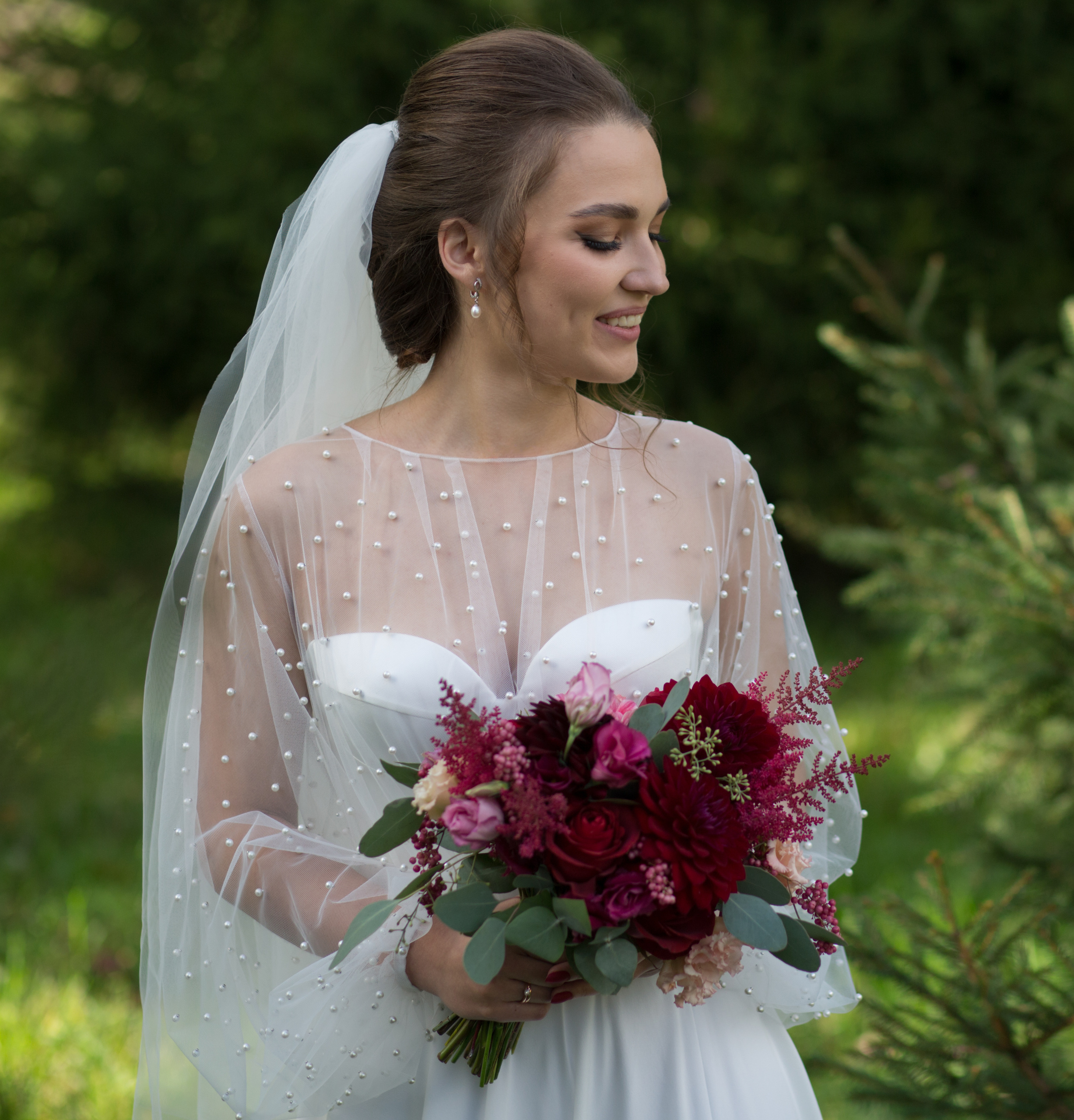 невеста смотрит в сторону, держа в руках красный букет на фоне зеленых деревьев в парке мытищи