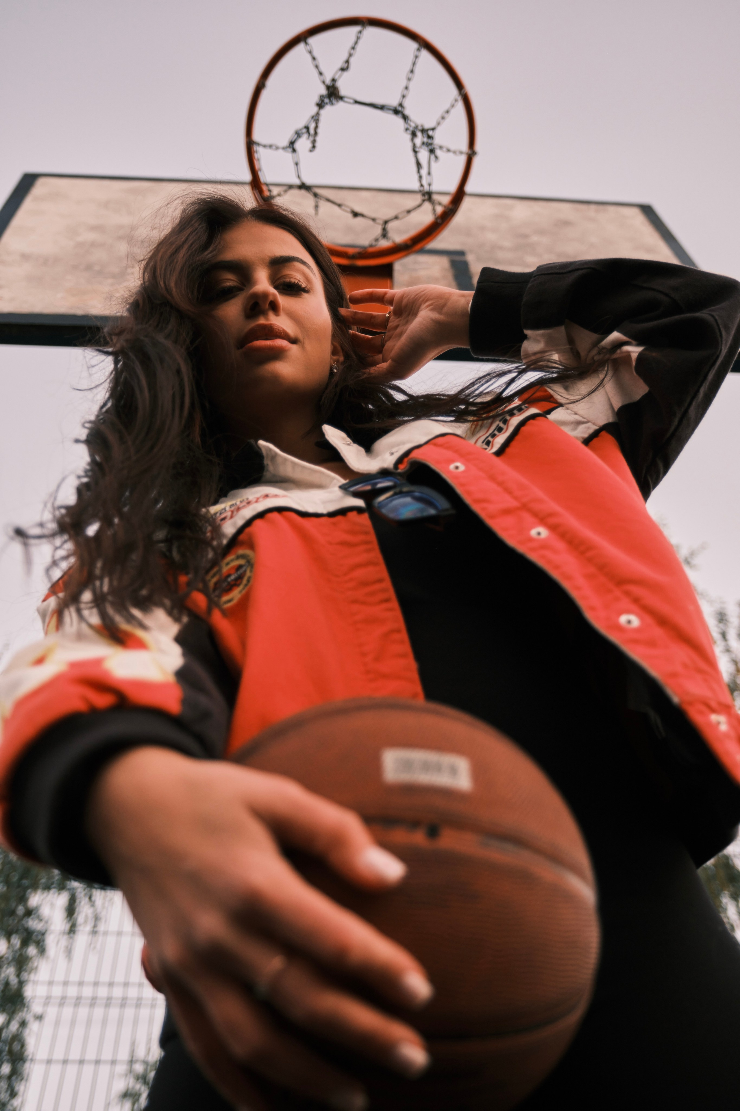 Кадр снизу вверх: девушка с мячом в руках стоит прямо под уличным баскетбольным кольцом.