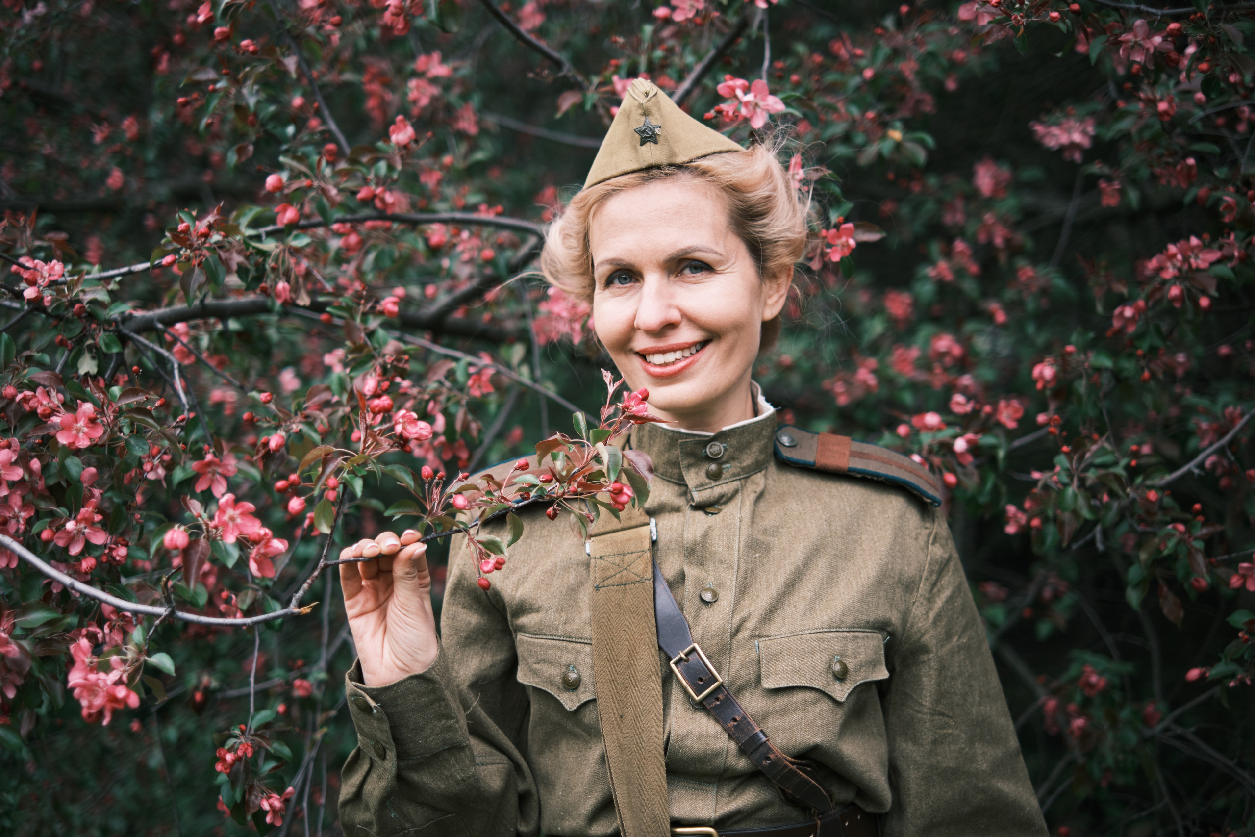 Девушка в военной форме и пилотке стоит на фоне цветущих яблонь