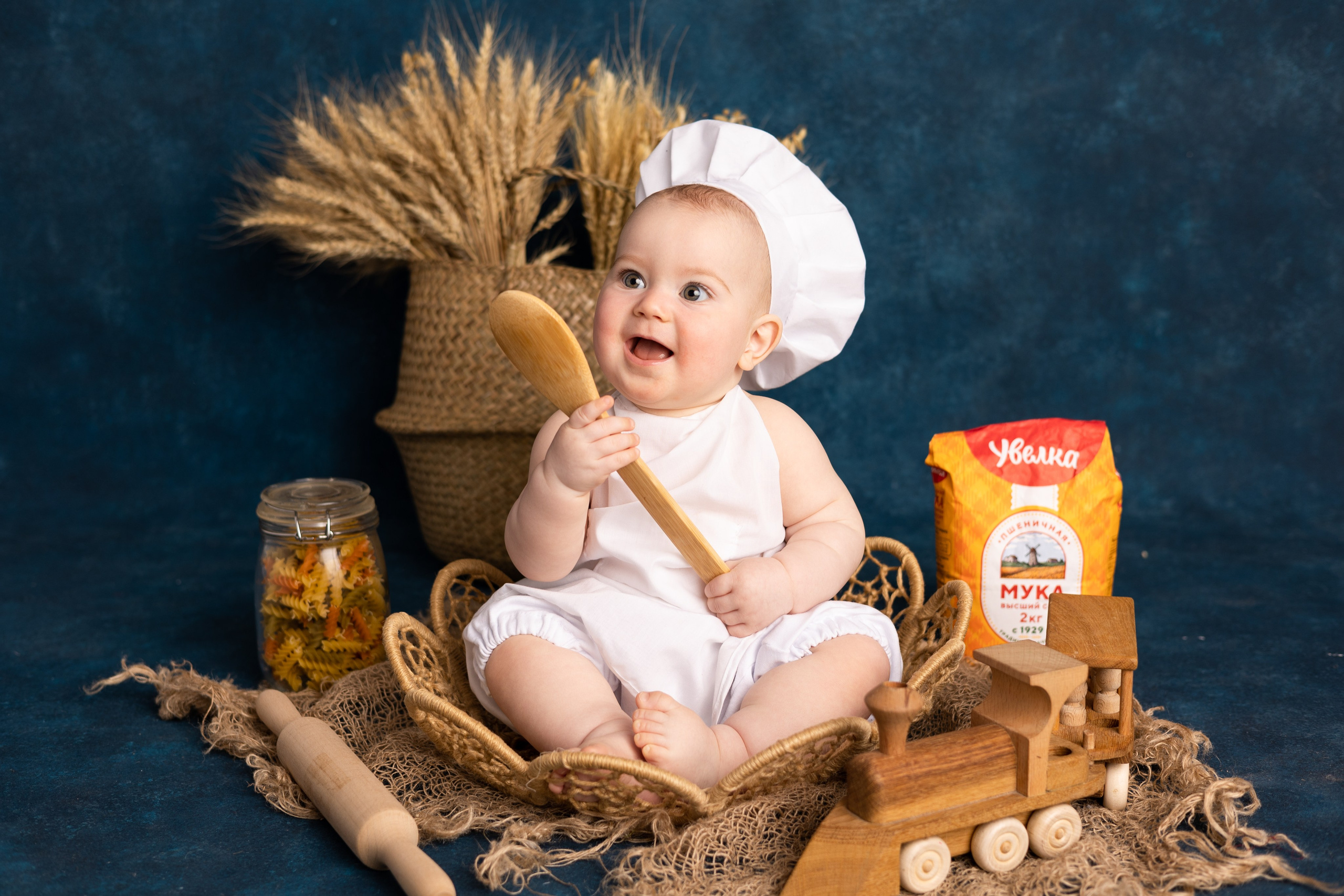 Счастливый младенец в костюме повара с деревянной лопаткой и продуктами на синем фоне.