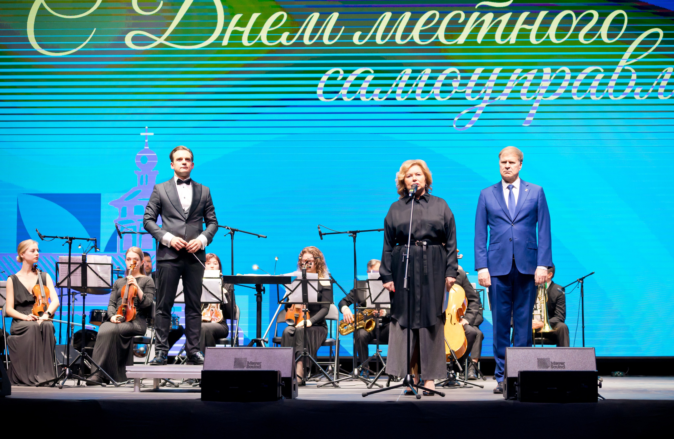 Торжественная часть мероприятия: женщина в чёрном выступает, рядом мужчина в синем костюме. На экране надпись «С Днём местного самоуправления». Фото Евгения Мальцева.