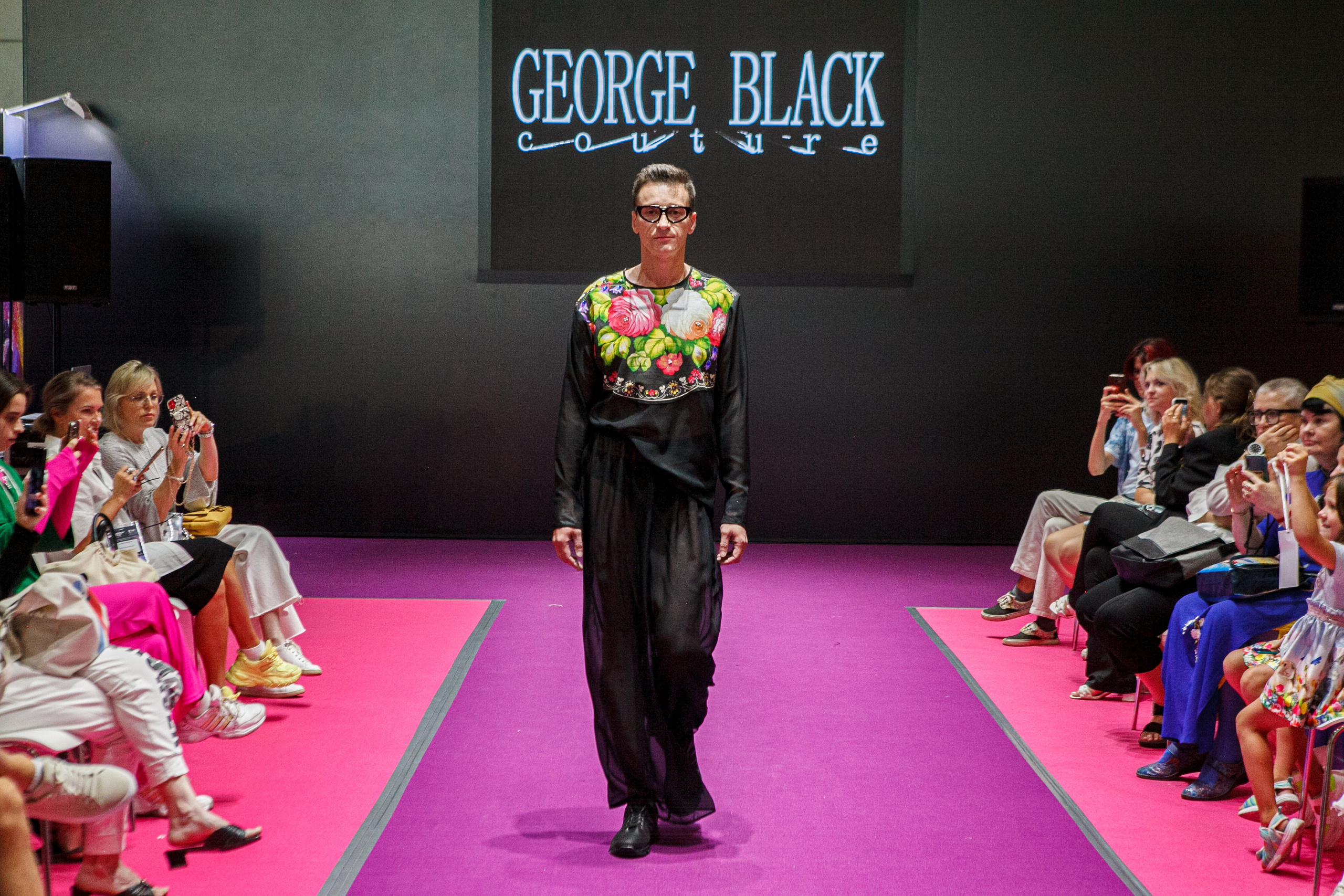  "Модели в ярких нарядах проходят по подиуму во время показа мод, освещенные яркими софитами и окруженные восторженными зрителями."