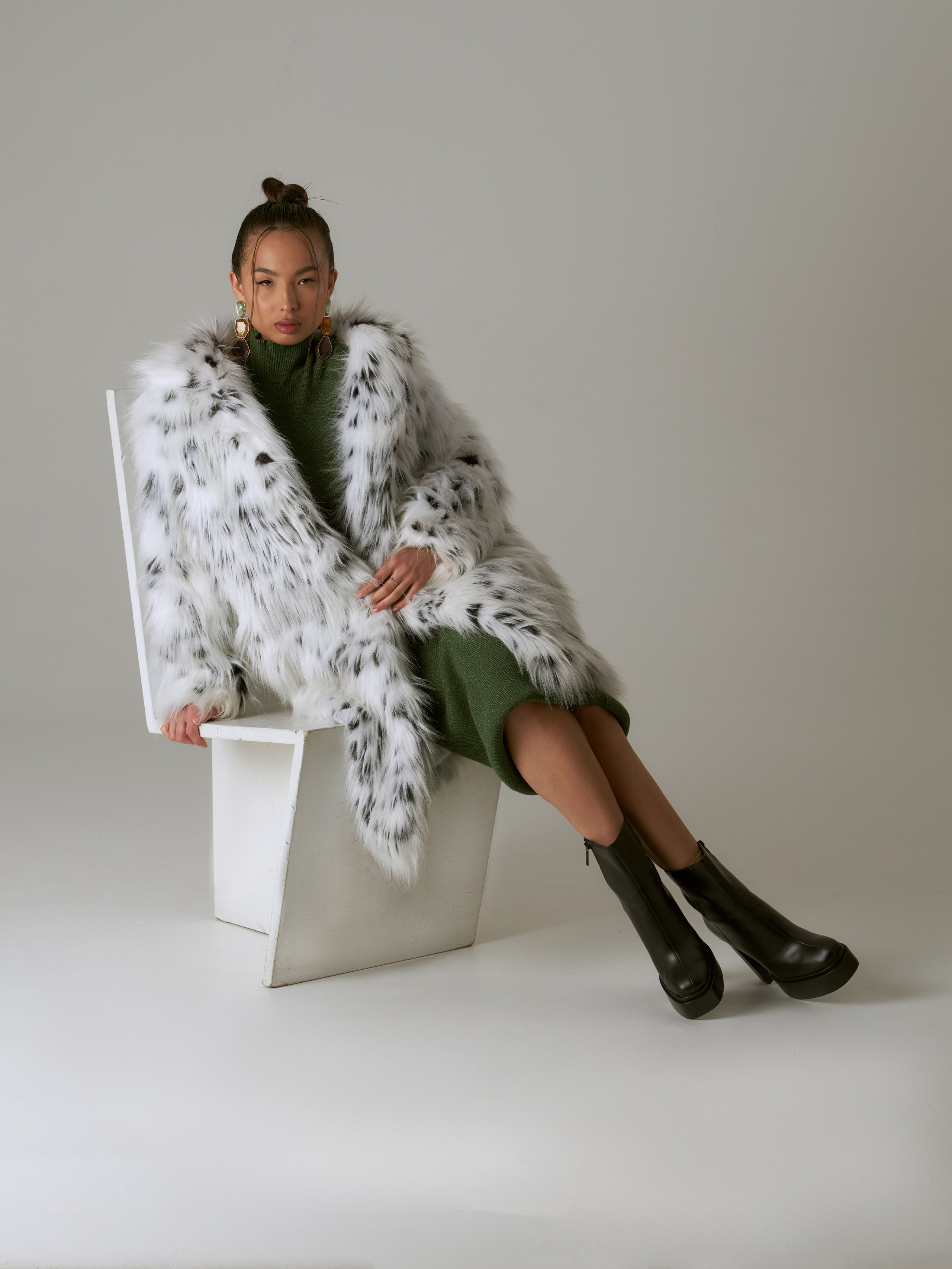 Девушка в зеленом платье и в шубе сидит на белом стуле на белой циклораме