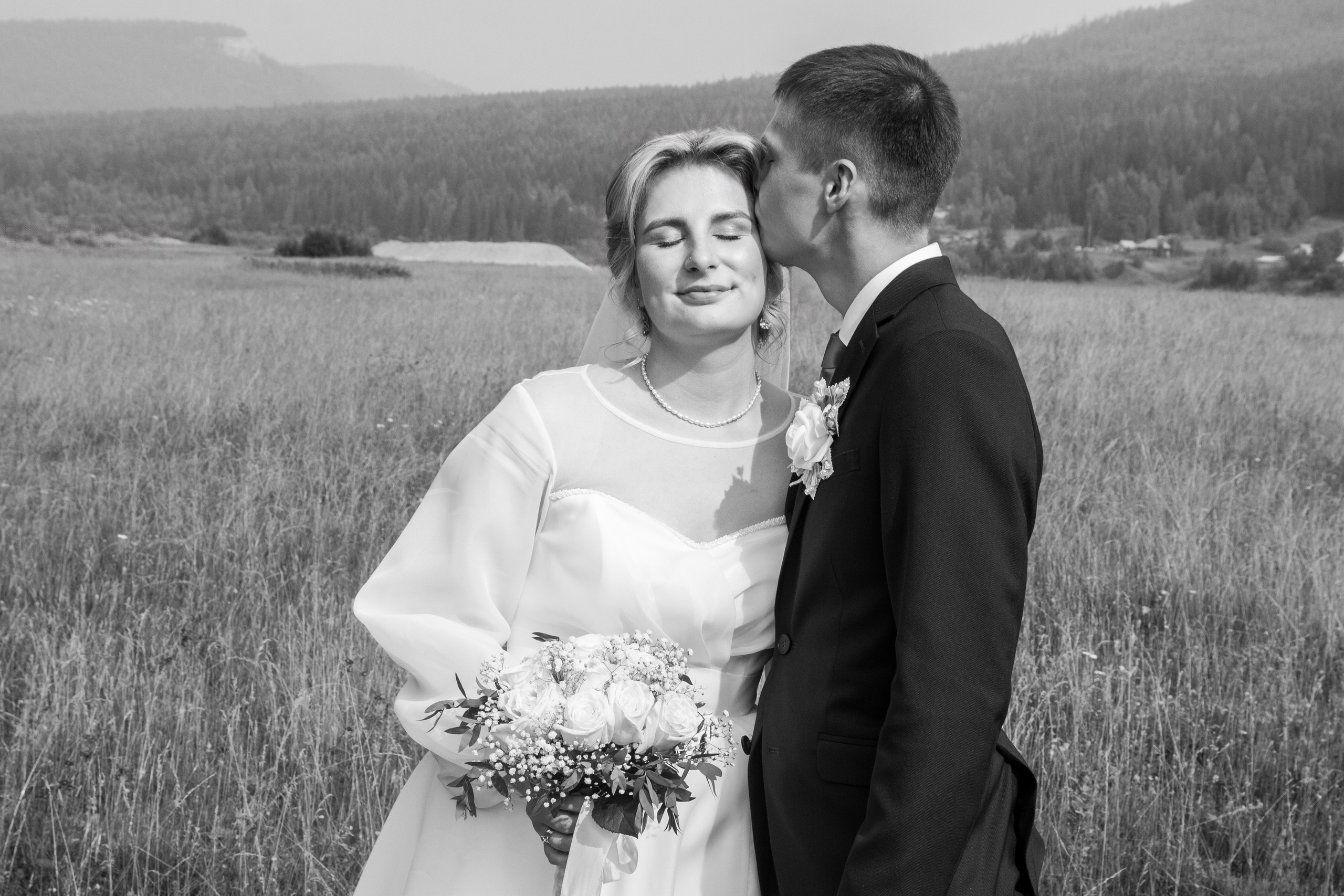 Чёрно-белый снимок: невеста улыбается, жених целует её в висок на фоне поля и холмов.