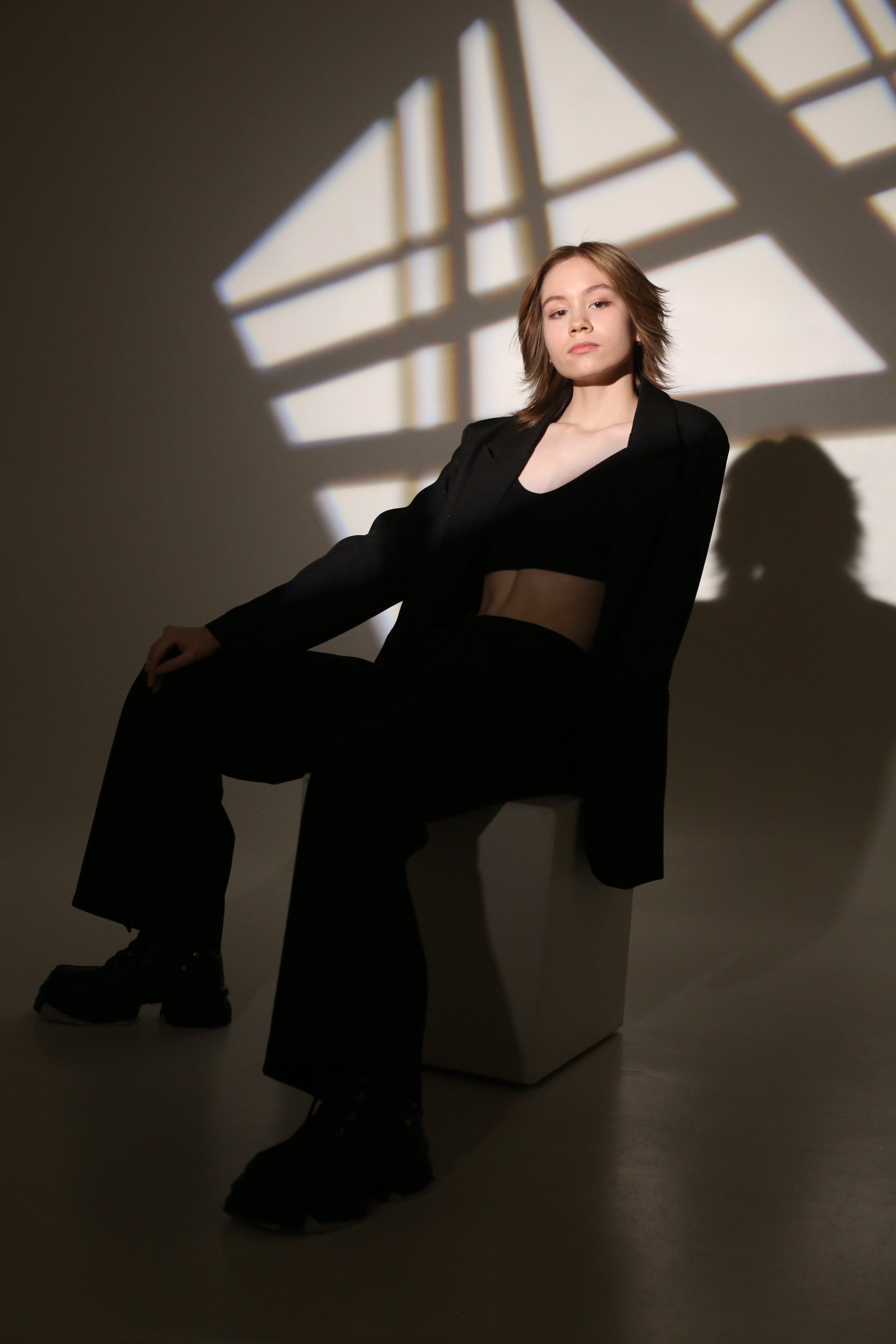Портрет из фотосессии «Геометрия»: девушка в черном костюме сидит на кубе в лучах света