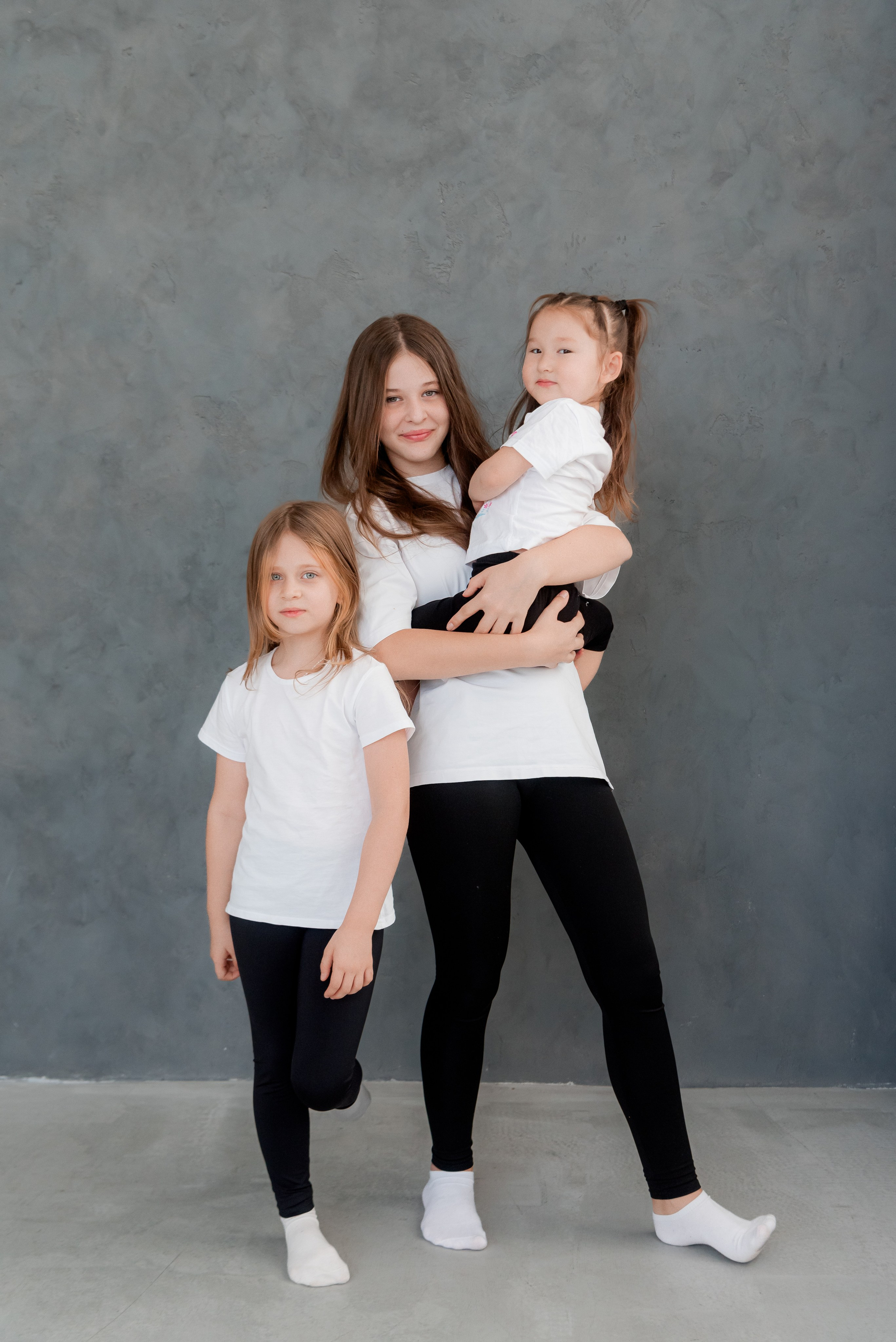 Три сестры обнимаются вместе старшие держит младшую на руках вместе одеты в один стиль белый верх футболки и чёрные Леггинсы