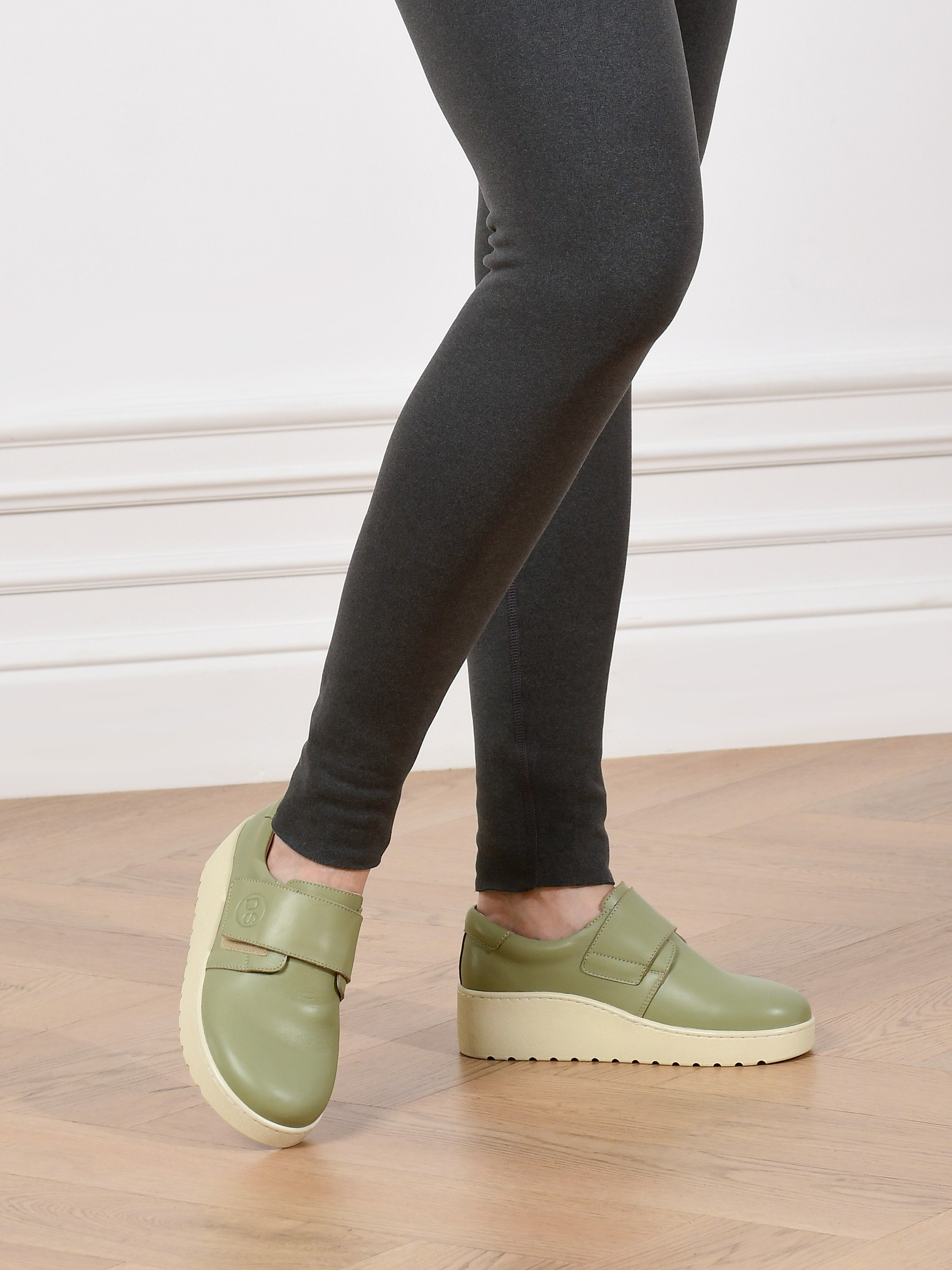 модель в зелёных туфлях на светлой подошве стоит на деревянном полу у белой стены.