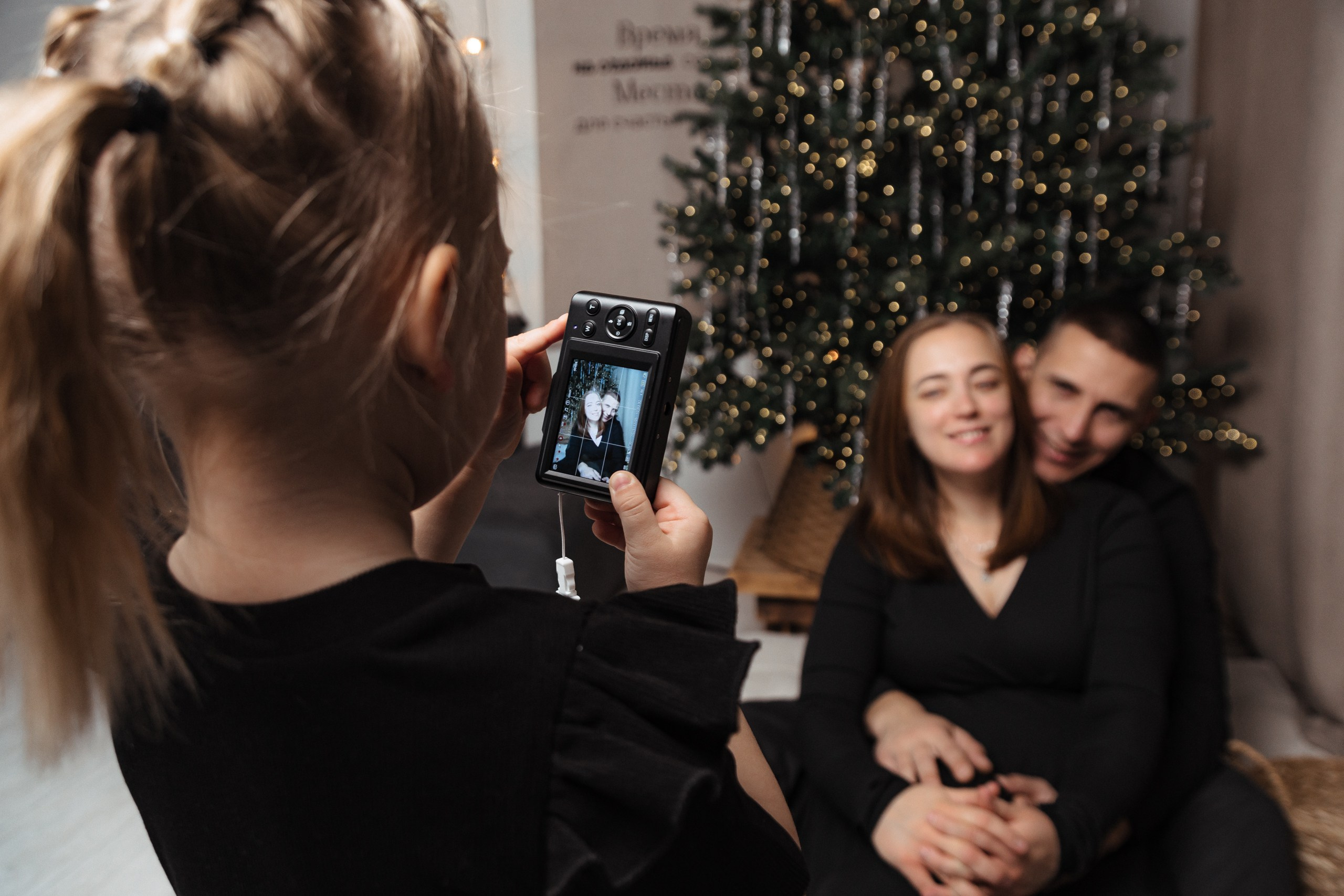 Вид со спины ребёнка, который фотографирует родителей на телефон у ёлки; момент семейного фото за кадром.
