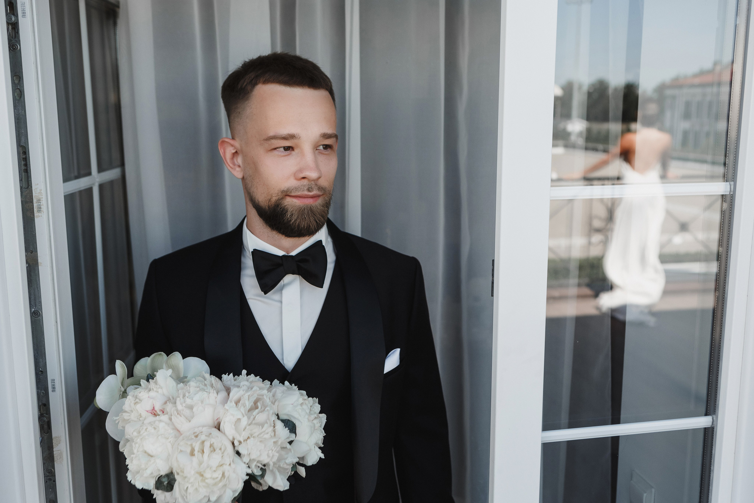 Жених с букетом у балконной двери, в отражении — силуэт невесты на террасе комплекса Golden Horse, свадебный фотограф Таганрог Антон Бакарюк