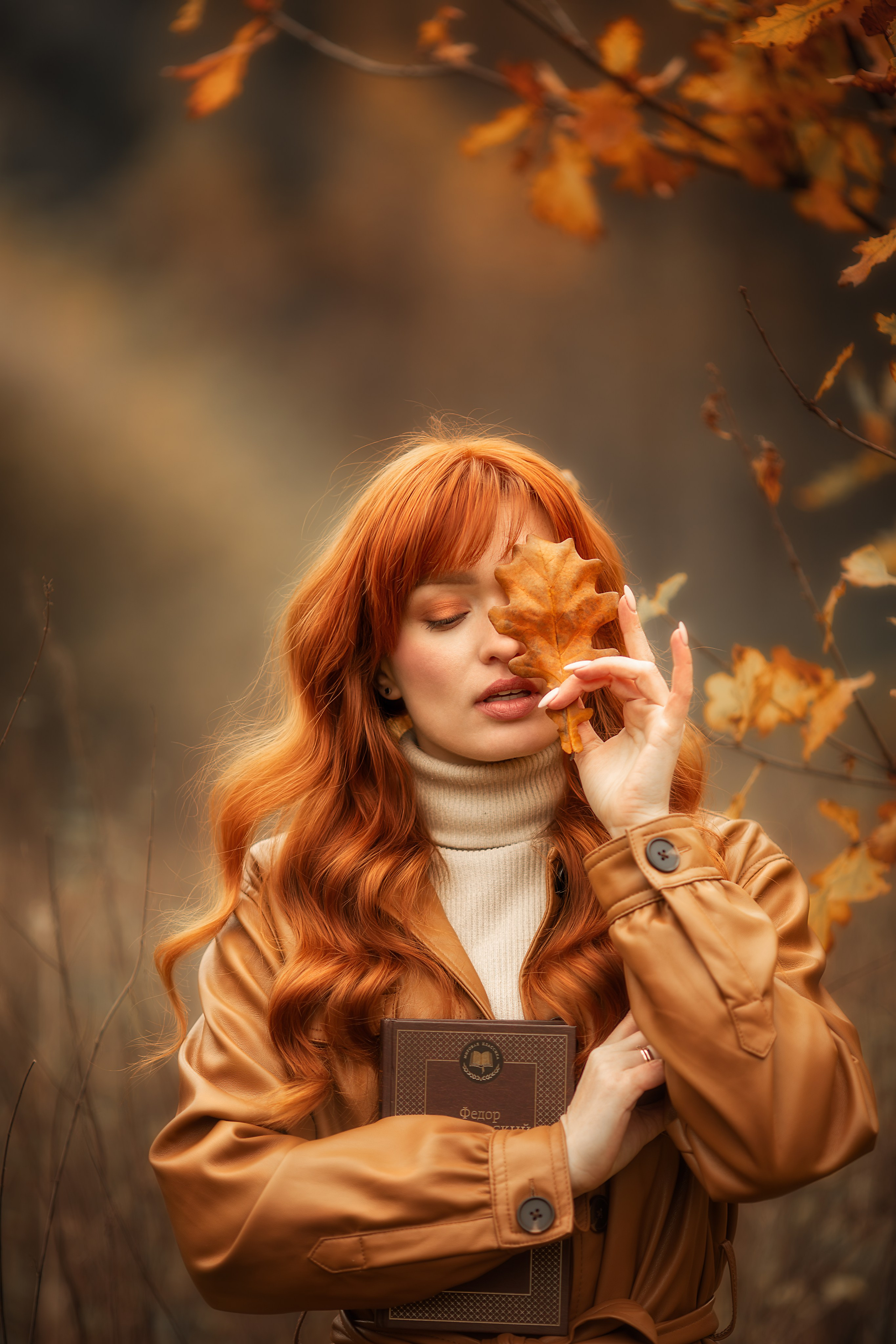 Девушка с рыжими волосами и осенним листом у лица, держит книгу.