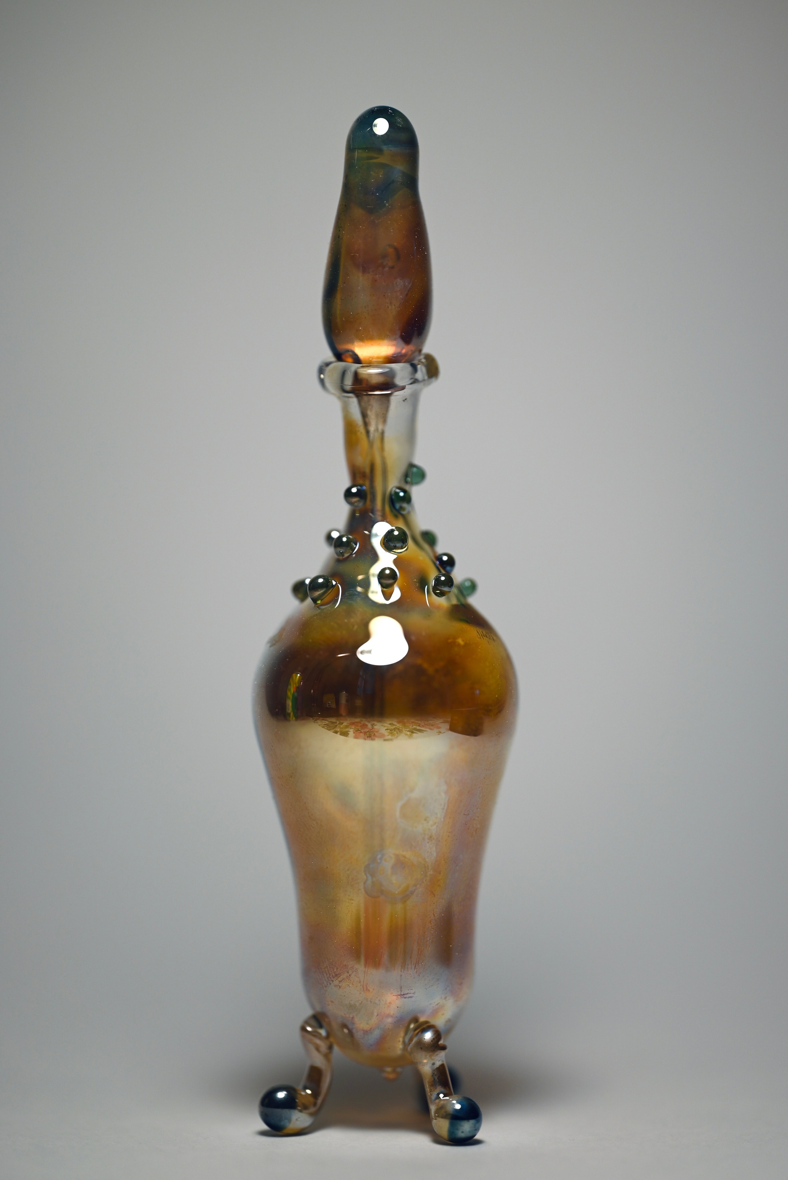 alt="Декоративная стеклянная скульптура с каплевидной крышкой, шарообразными ножками и округлым телом с янтарно-золотистыми переливами и тёмными бусинами у основания горлышка."