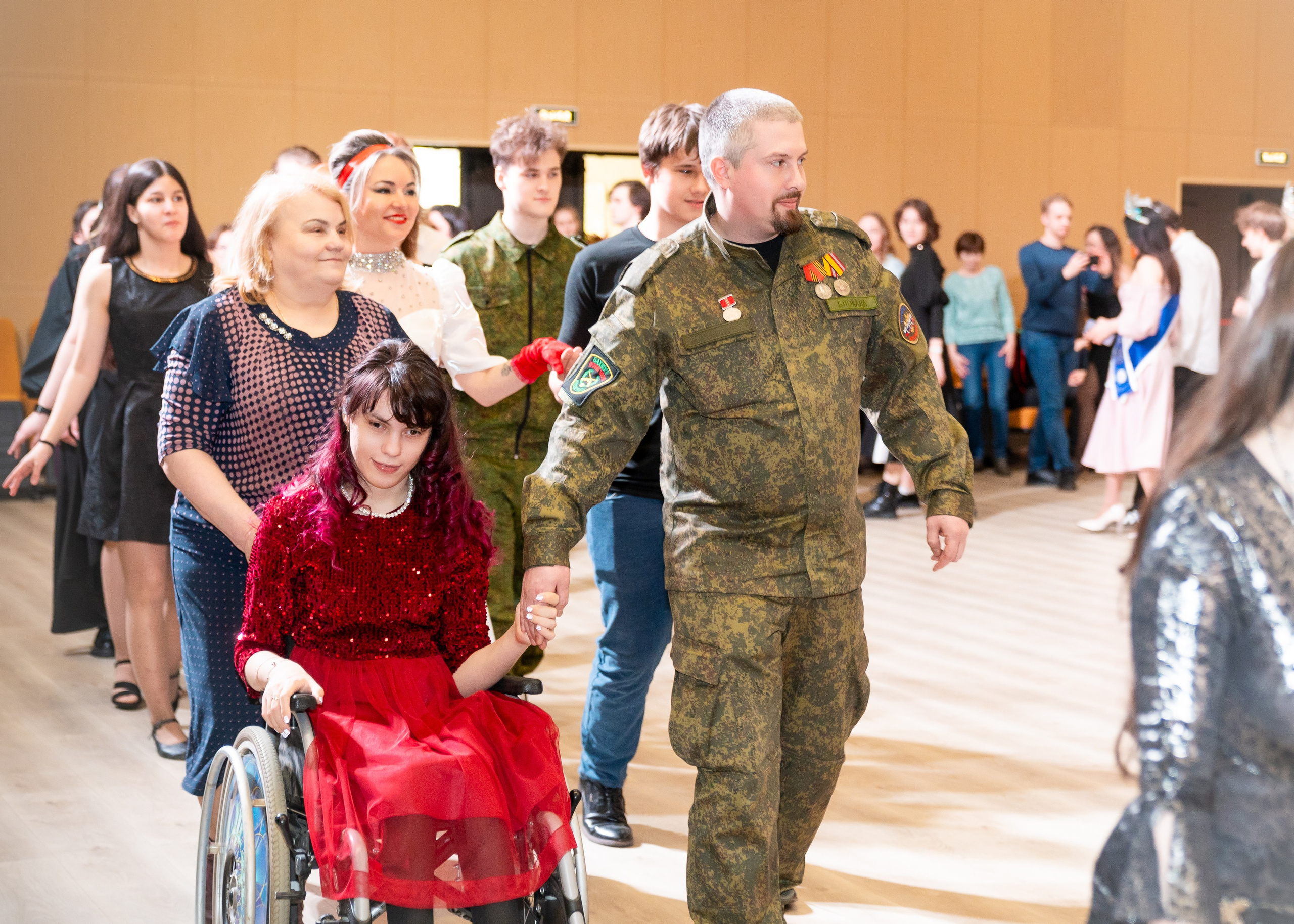 Мужчина в камуфляжной форме держит за руку девушку в красном платье на инвалидной коляске на инклюзивном балу проекта «Мы все равны». Фотограф Мальцев Евгений.